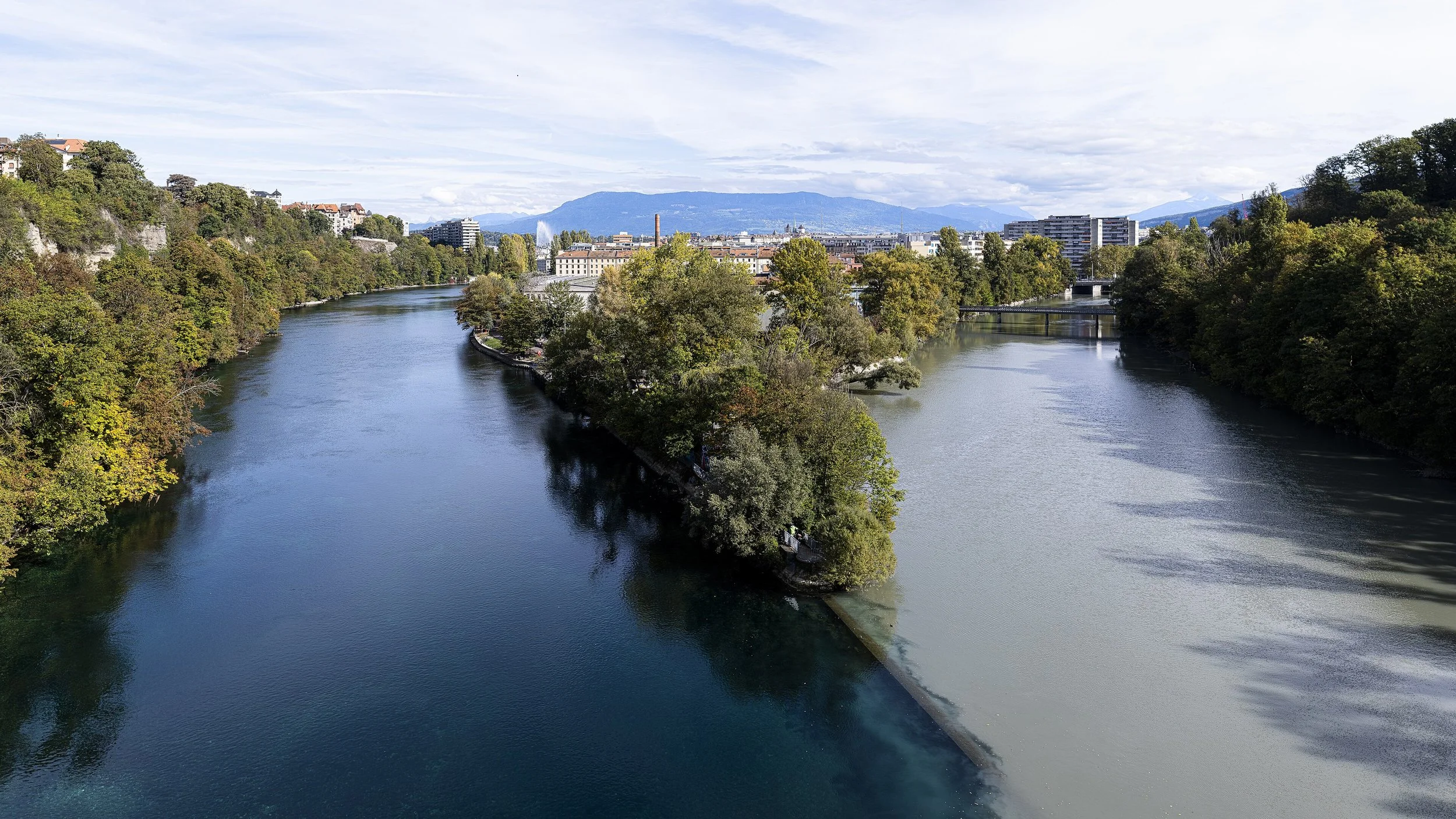 Vue d'une rivière serpentant à travers une ville avec des bâtiments modernes au fond, entourée d'arbres verts et de montagnes en arrière-plan, sous un ciel partiellement nuageux.