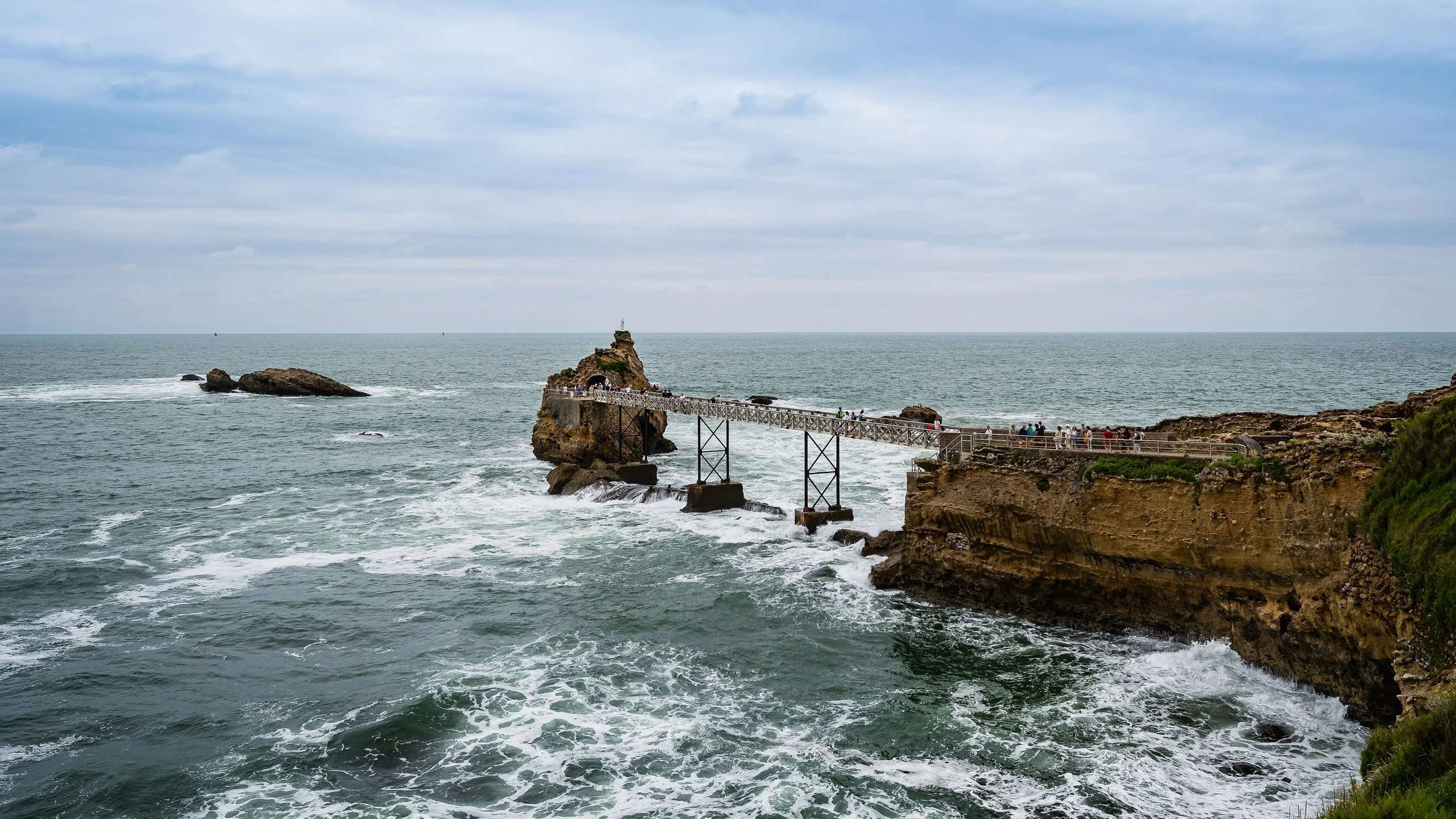 Une jetée en bois reliant la côte à un rocher sur l'océan, avec des gens qui marchent dessus, sous un ciel partiellement nuageux.