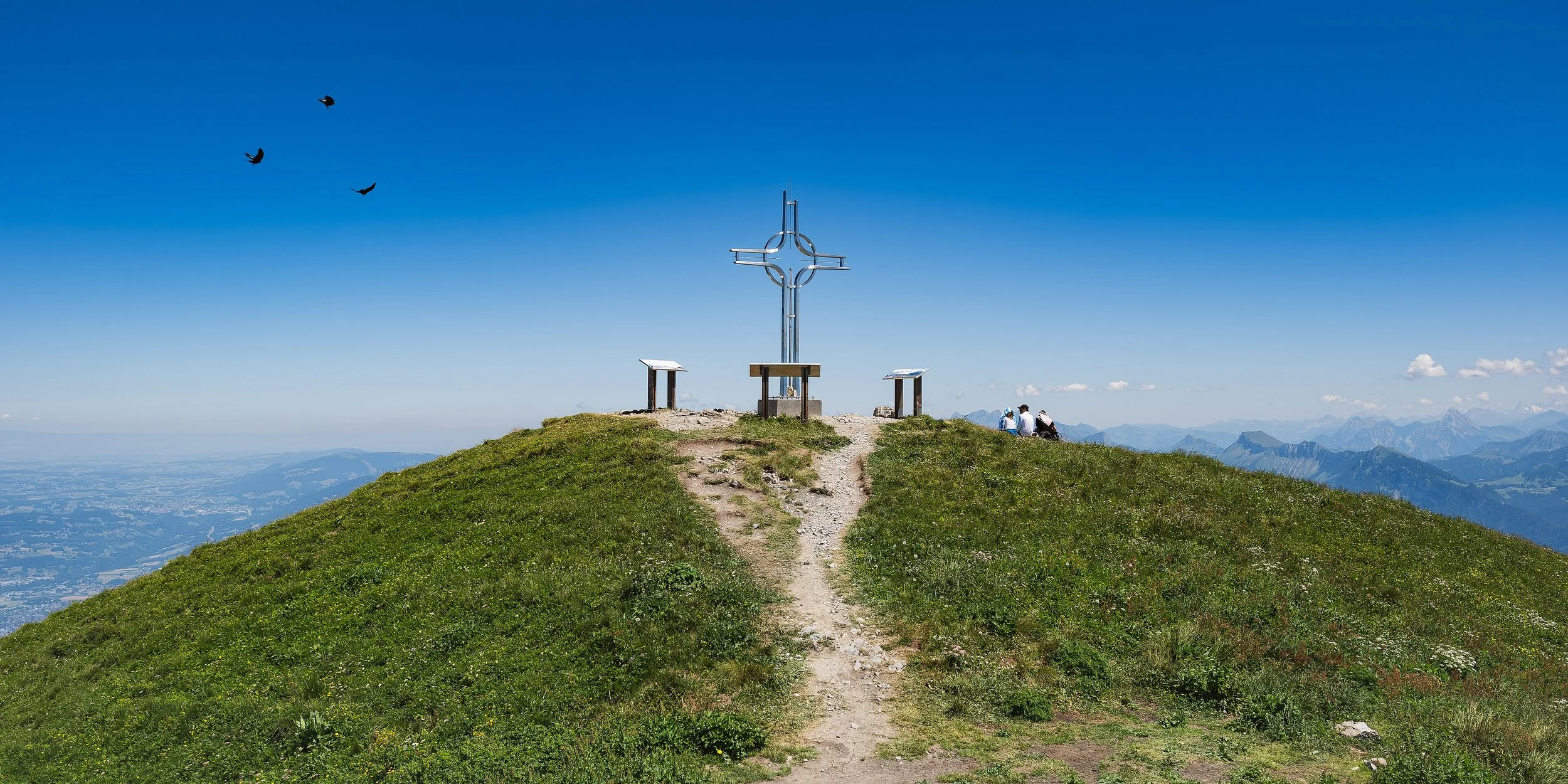 Montagne avec un sommet en herbe, trois panneaux d'information, un grand croix métallique, et deux personnes assises regardant la vue, sous un ciel bleu avec quelques nuages, vue sur des montagnes en arrière-plan.