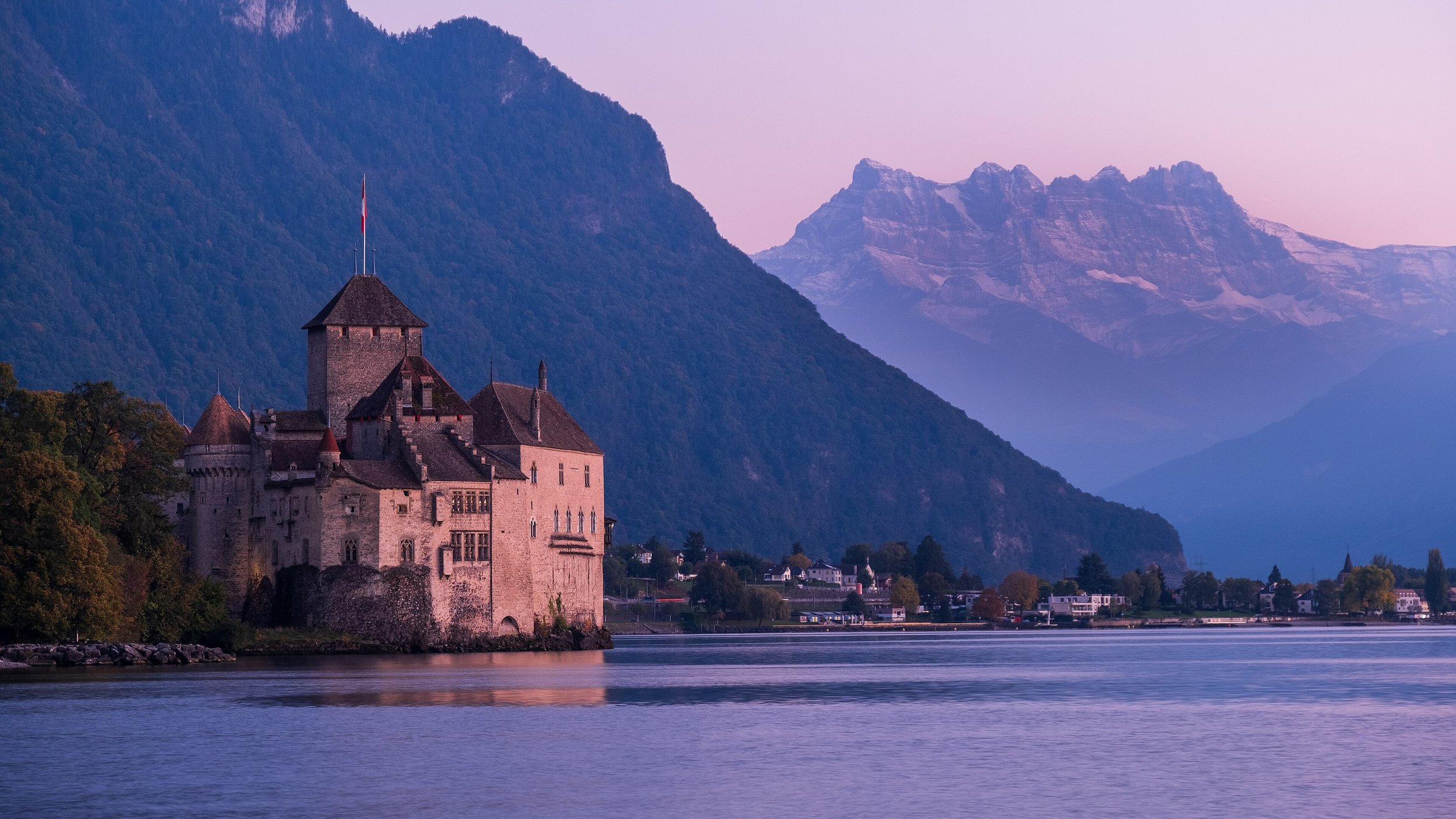Un château médiéval situé au bord d'une rivière, avec des montagnes en arrière-plan au crépuscule.