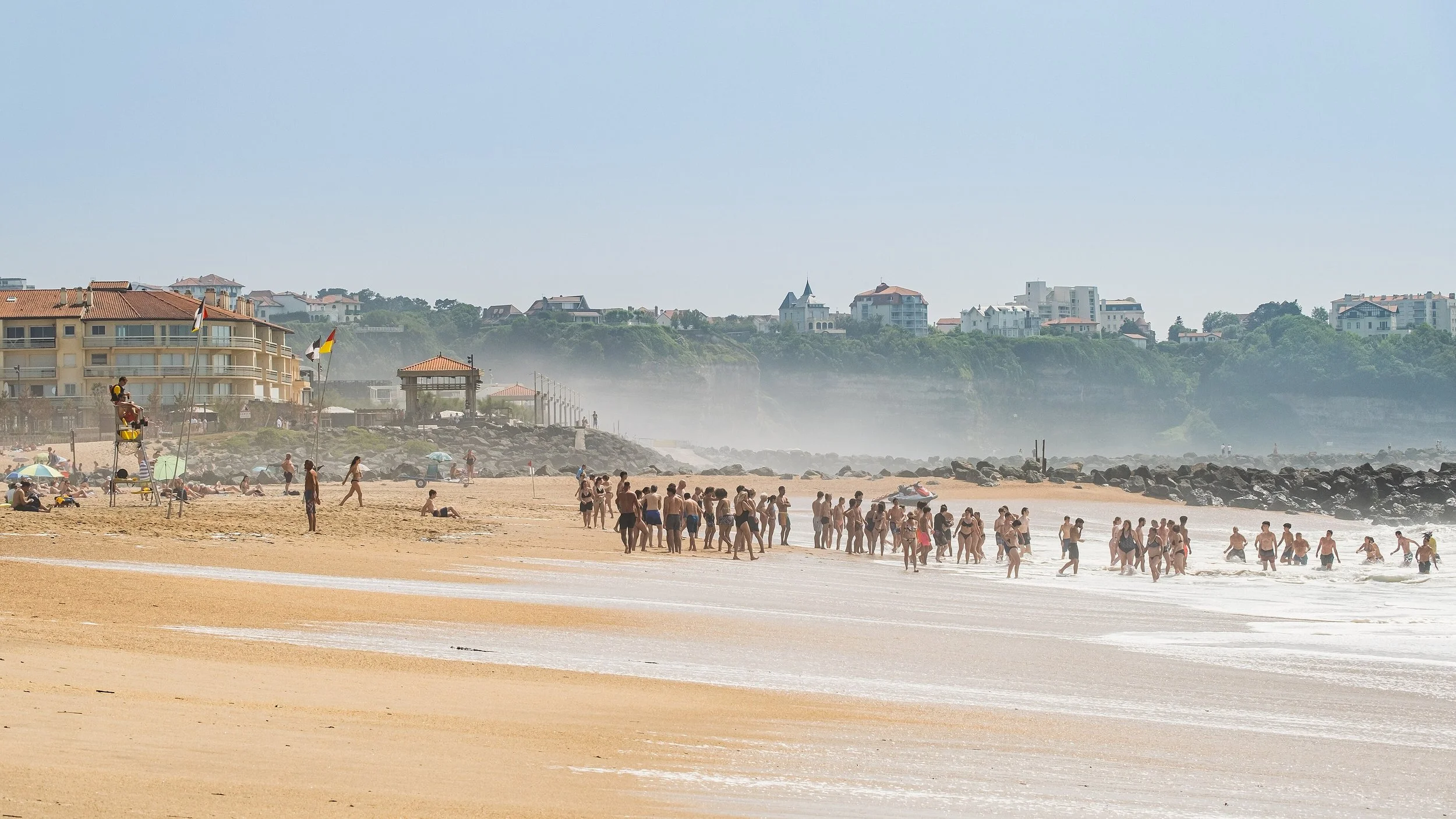 Plage bondée avec des gens nageant et se relaxant, bâtiments résidentiels en arrière-plan, brume marine et ciel clair.