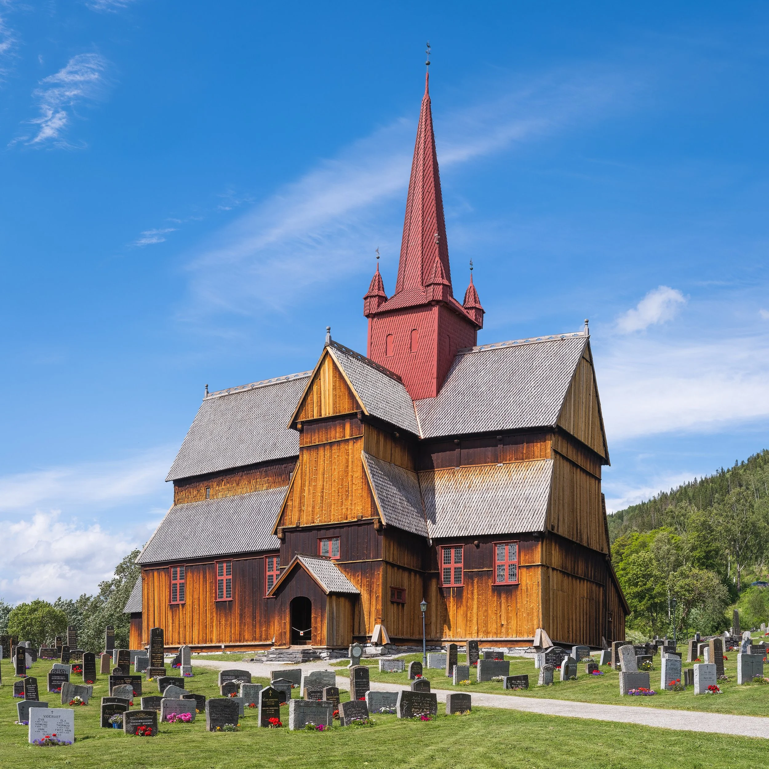 Une église en bois rouge avec un haut clocher pointu, située dans un cimetière avec de nombreuses tombes, sous un ciel bleu clair avec quelques nuages.
