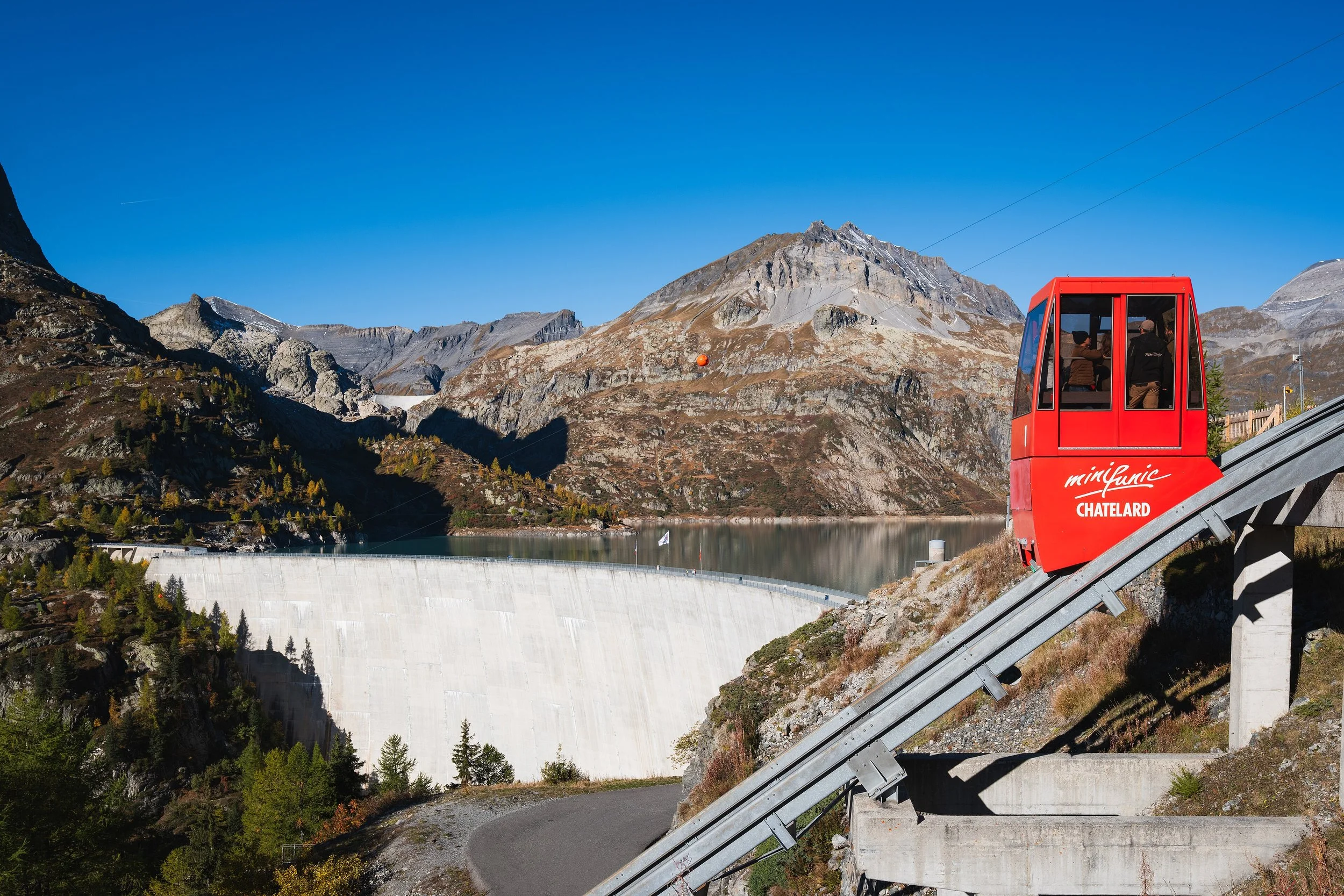 Téléphérique rouge avec deux personnes à l'intérieur, en montagne avec un lac et un barrage derrière, et des montagnes rocheuses en arrière-plan, sous un ciel bleu clair.