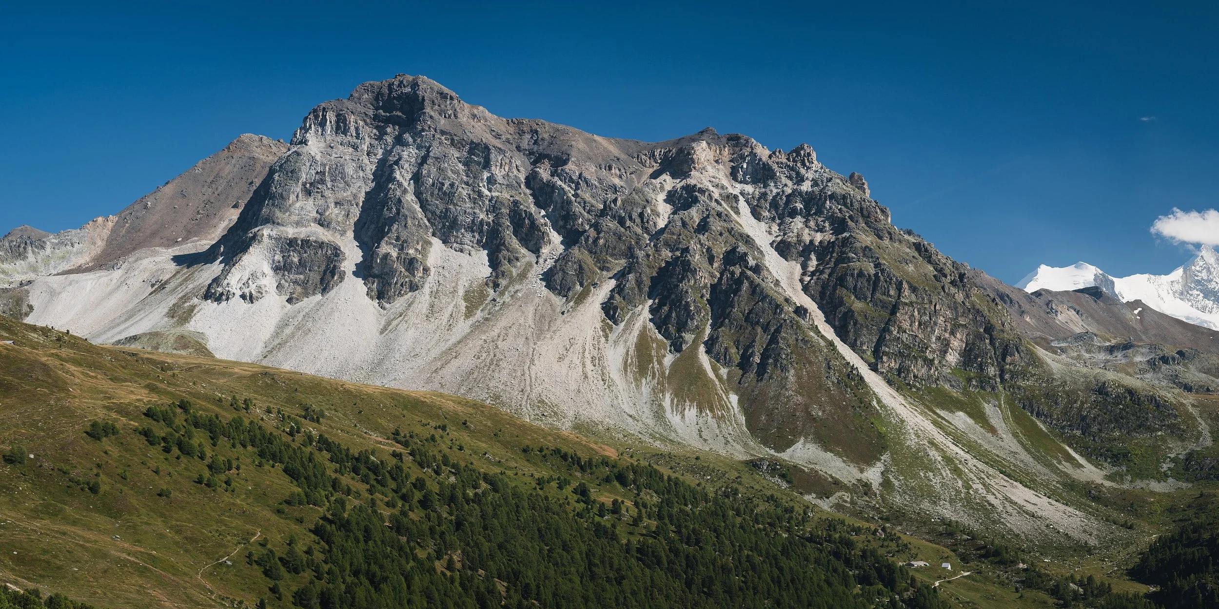 Montagne escarpée avec des pentes rocheuses et des zones de végétation verte au premier plan, ciel bleu clair.