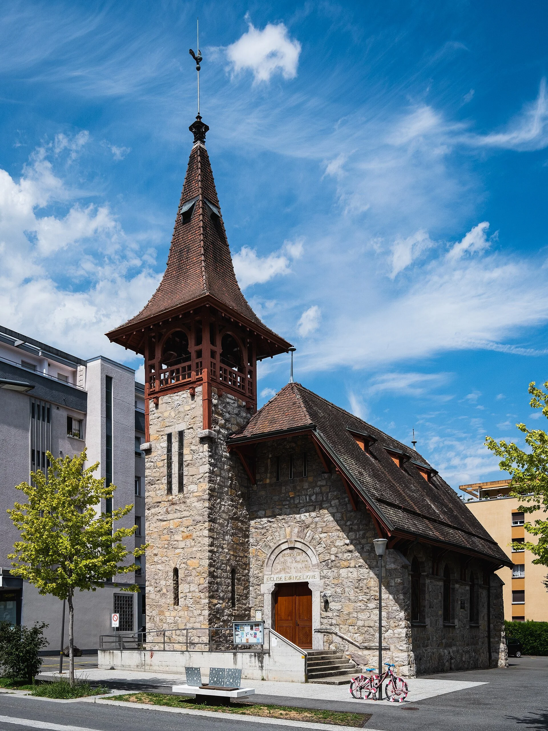 Une petite église en pierre avec un clocher en bois et un toit en tuiles rouges, située dans un environnement urbain avec des bâtiments modernes en arrière-plan. Il y a deux vélos décorés en face de l’église et un banc en béton à proximité.