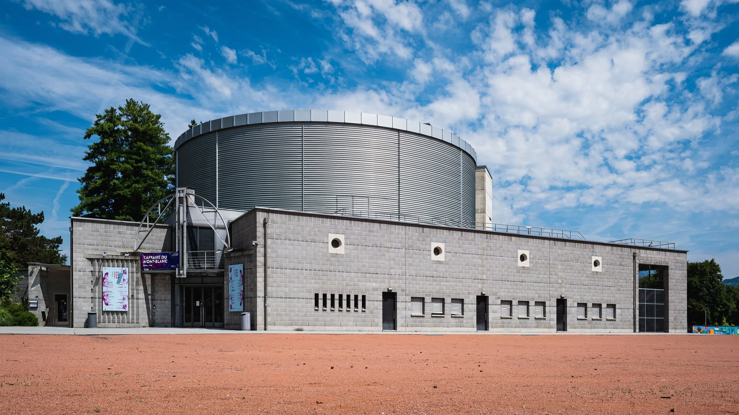 Un bâtiment moderne avec une structure en béton et une grande coupole métallique au sommet, entouré d'un espace ouvert avec un sol en terre battue et un ciel bleu avec quelques nuages.