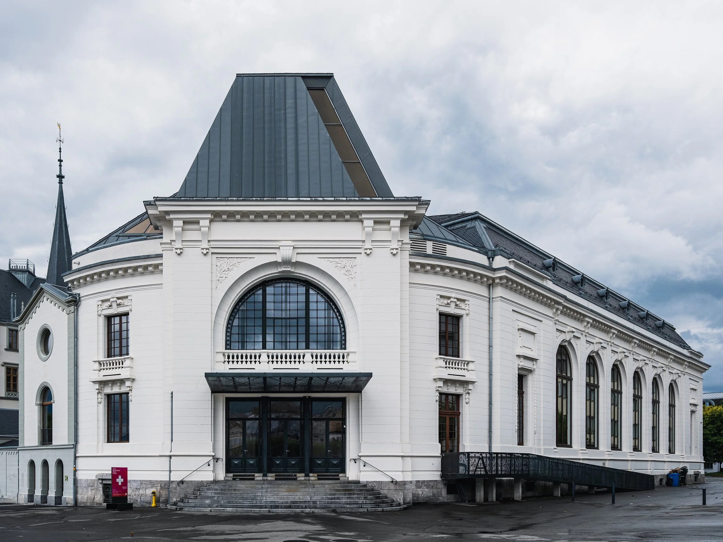 Bâtiment blanc à architecture classique avec grandes fenêtres et une entrée principale en acier, entouré de marches et une petite rampe d'accès, sous un ciel nuageux.