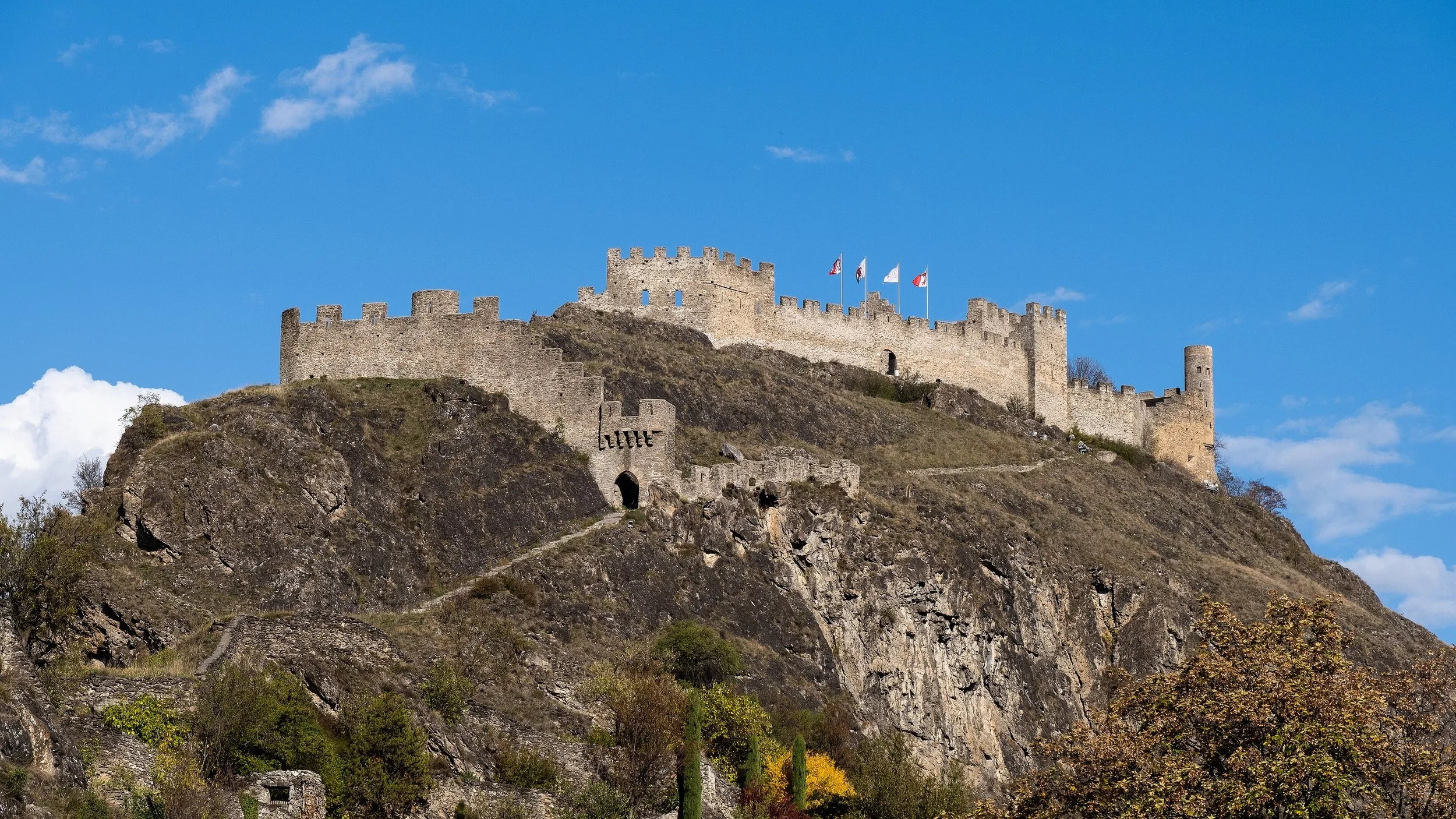 Château médiéval perché sur une colline rocheuse, avec des murs en pierre et des drapeaux flottant au sommet, sous un ciel bleu avec quelques nuages.