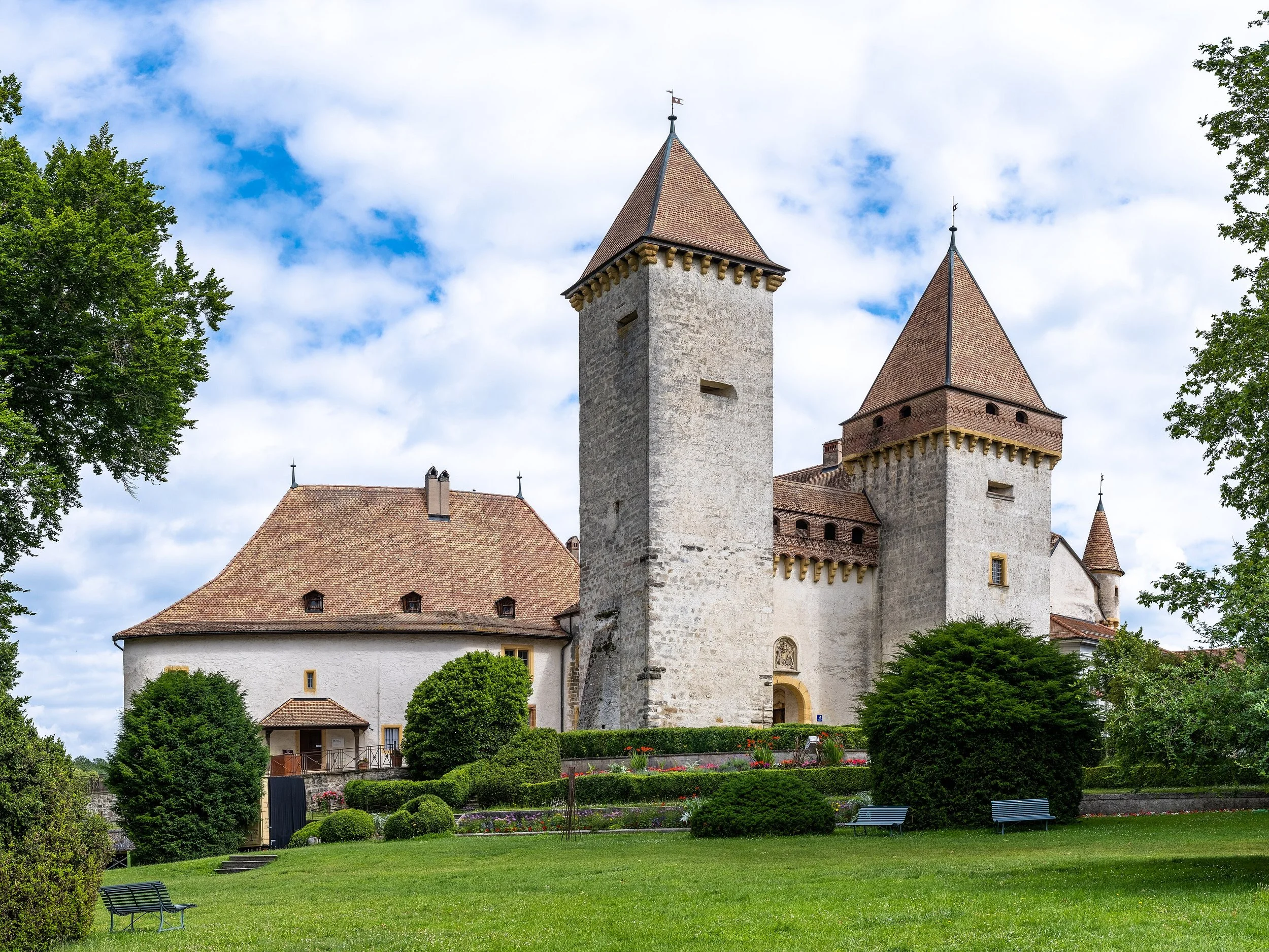 Une scène de château avec deux tours en pierre, entouré d'un jardin bien entretenu avec des buissons, des arbres et des bancs, sous un ciel partiellement nuageux.