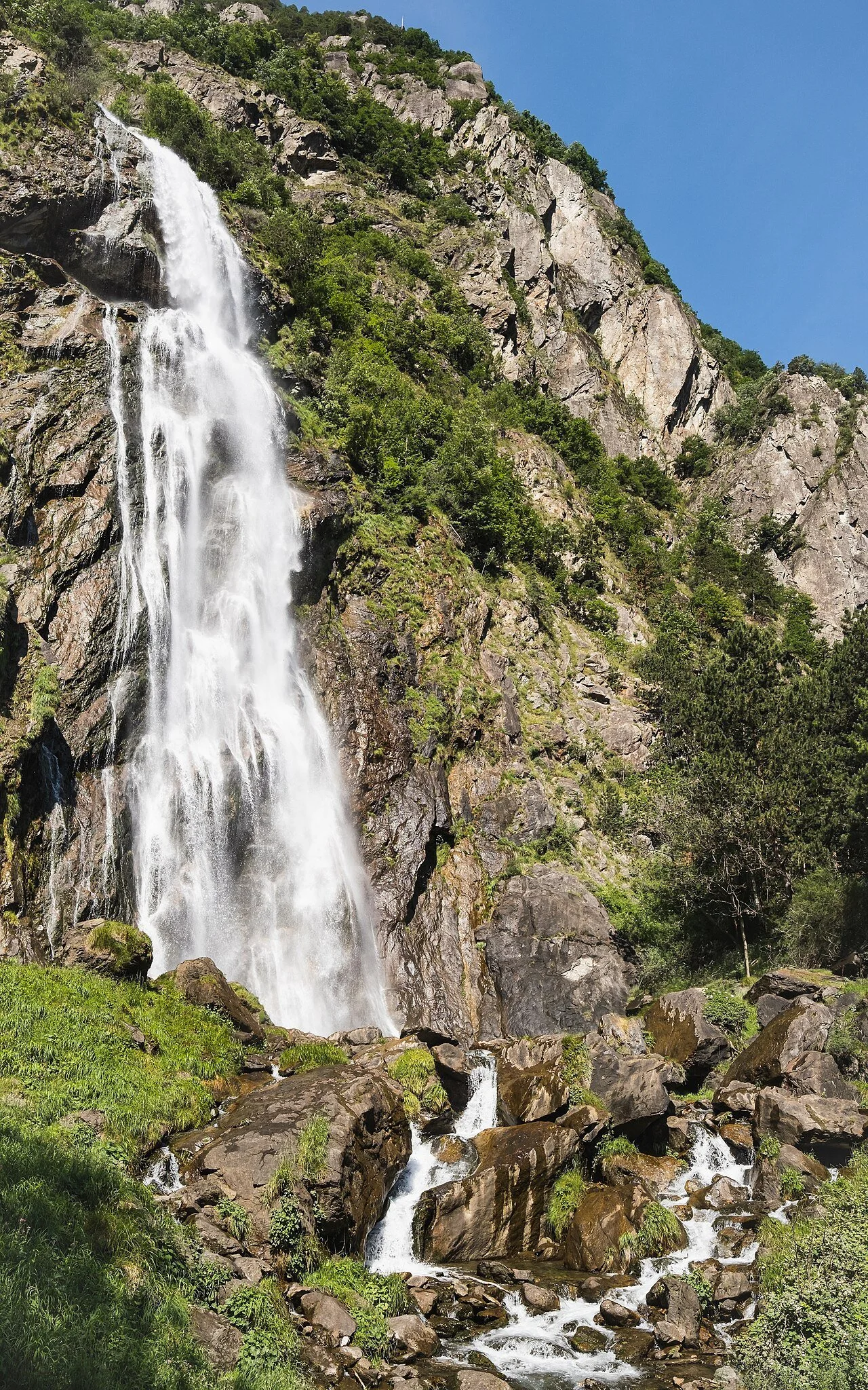 Cascade d'eau tombant d'une falaise rocheuse dans un environnement naturel verdoyant avec un ciel bleu.
