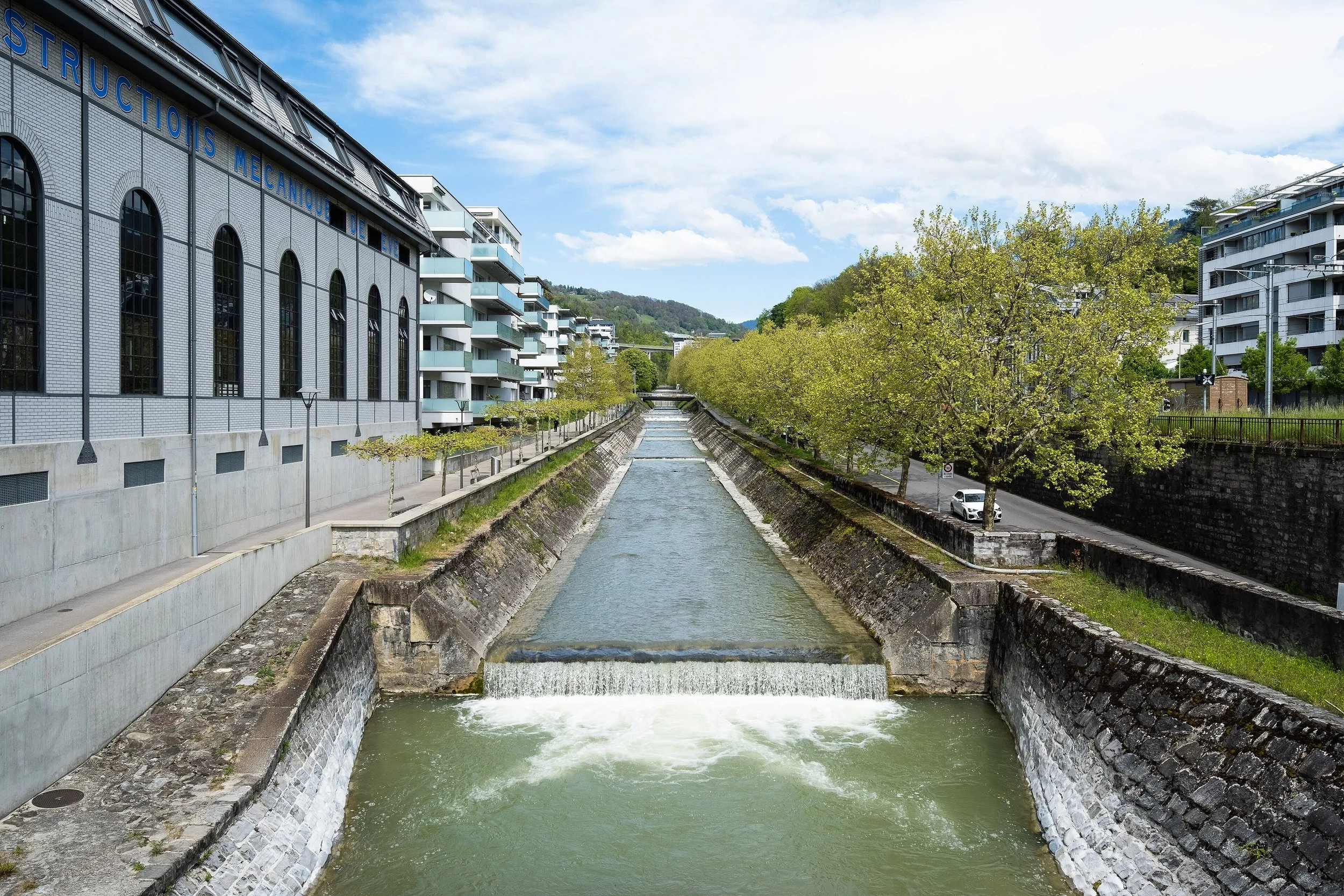 Une rivière urbaine traverse un quartier résidentiel avec des bâtiments modernes, des arbres verts et un ciel partiellement nuageux.