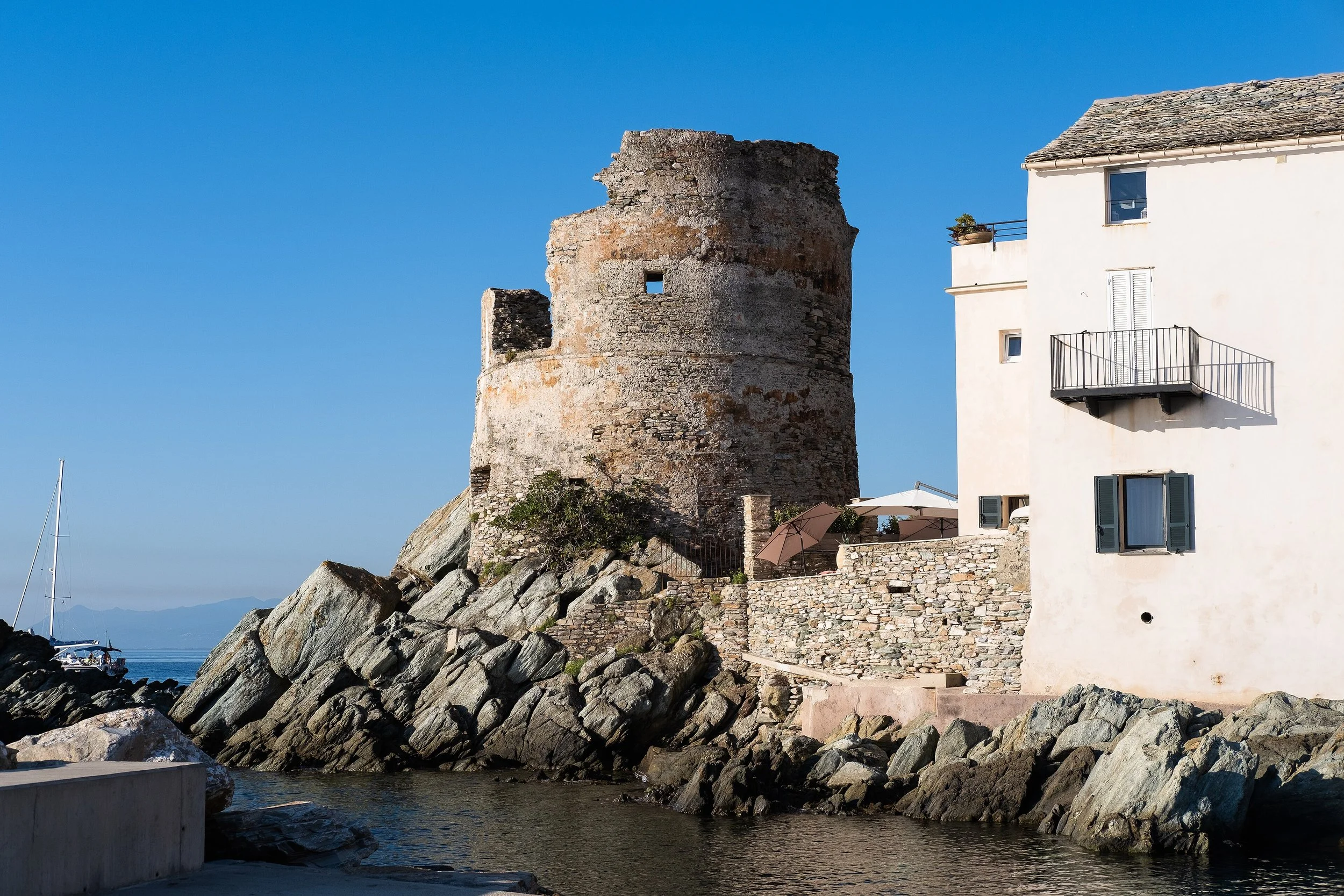 Tour en pierre ancienne à côté d'une maison blanche avec balcon sur la côte, entourée de rochers et d'eau, sous un ciel bleu clair.