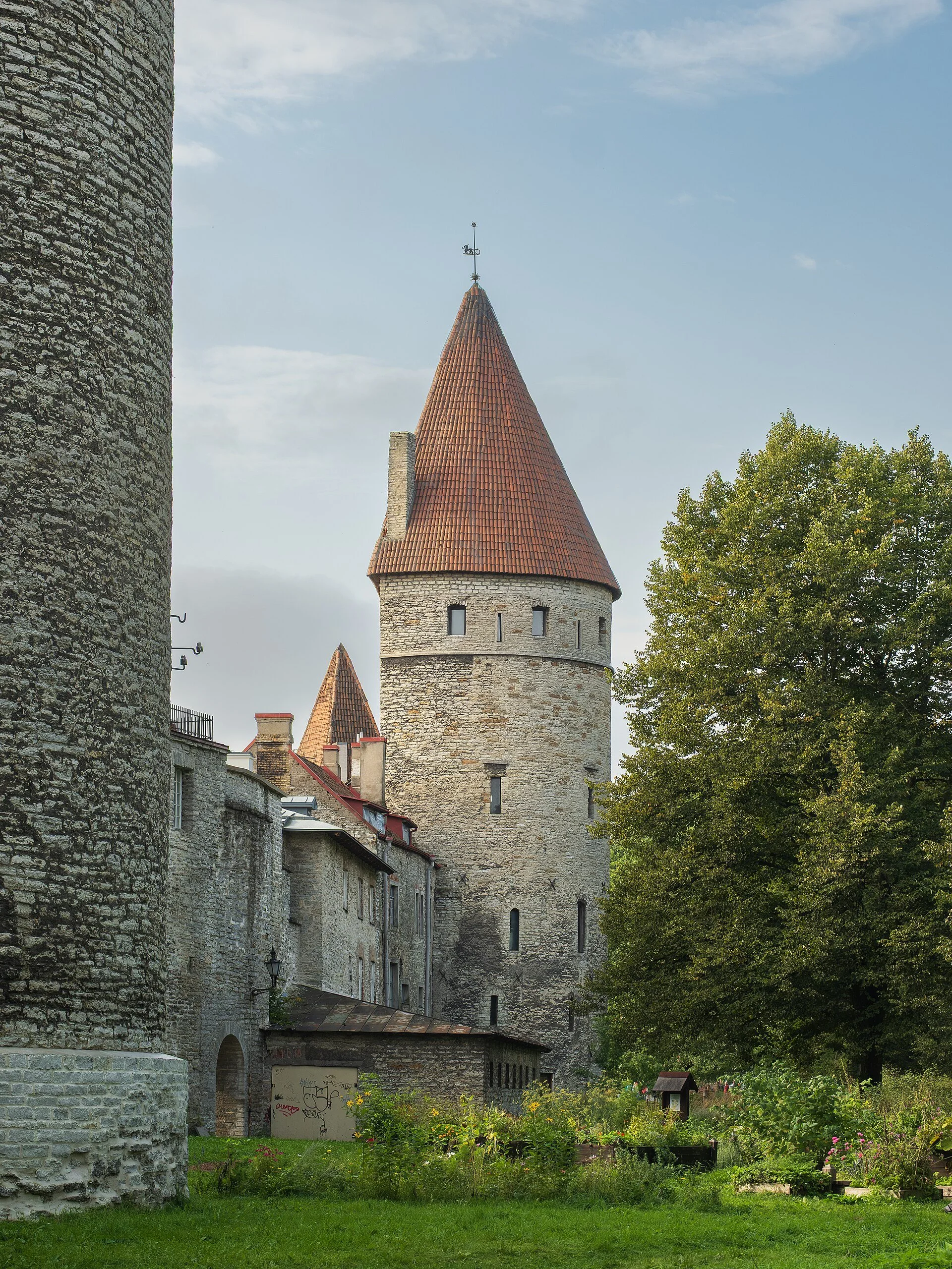 Vue d'une vieille tour médiévale en pierre avec un toit conique en tuiles rouges, entourée d'arbres et de bâtiments anciens en pierre, under un ciel partiellement nuageux.
