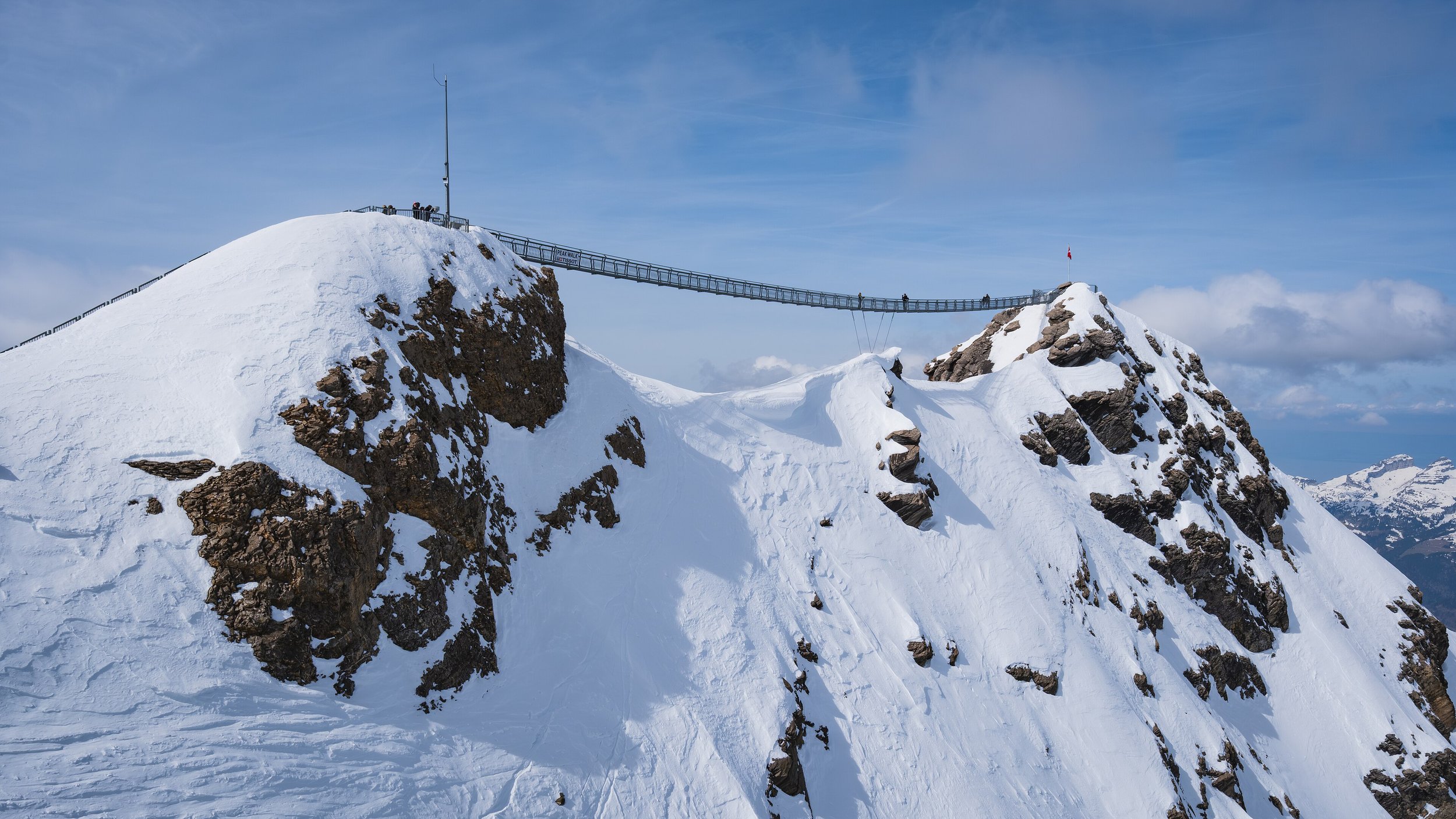 Un pont suspendu en métal traverse un sommet de montagne enneigé, avec un drapeau à une extrémité et une pancarte signalisant une randonnée.
