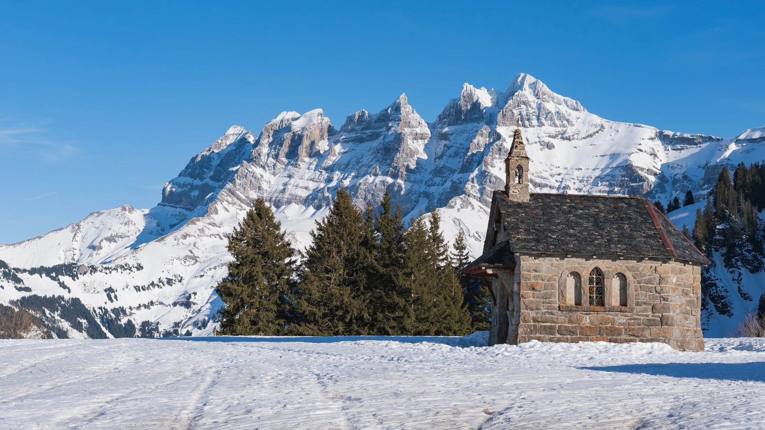Une petite église en pierre avec un toit en ardoise, située dans un paysage enneigé avec des montagnes couvertes de neige en arrière-plan, des arbres verts et un ciel bleu clair.
