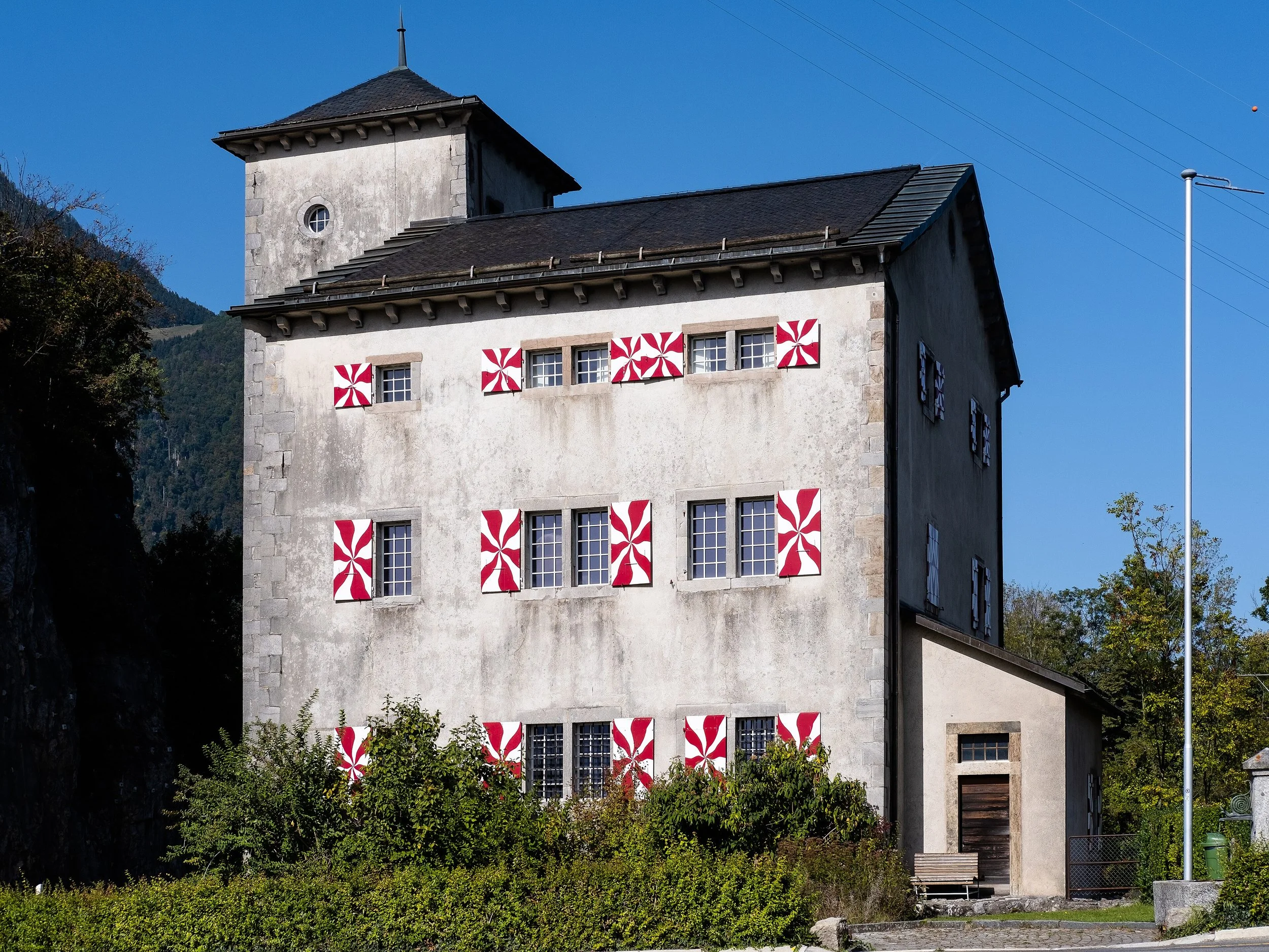 Une vieille maison en pierre avec des volets décorés de motifs rouges et blancs, située dans un environnement montagneux sous un ciel clair.