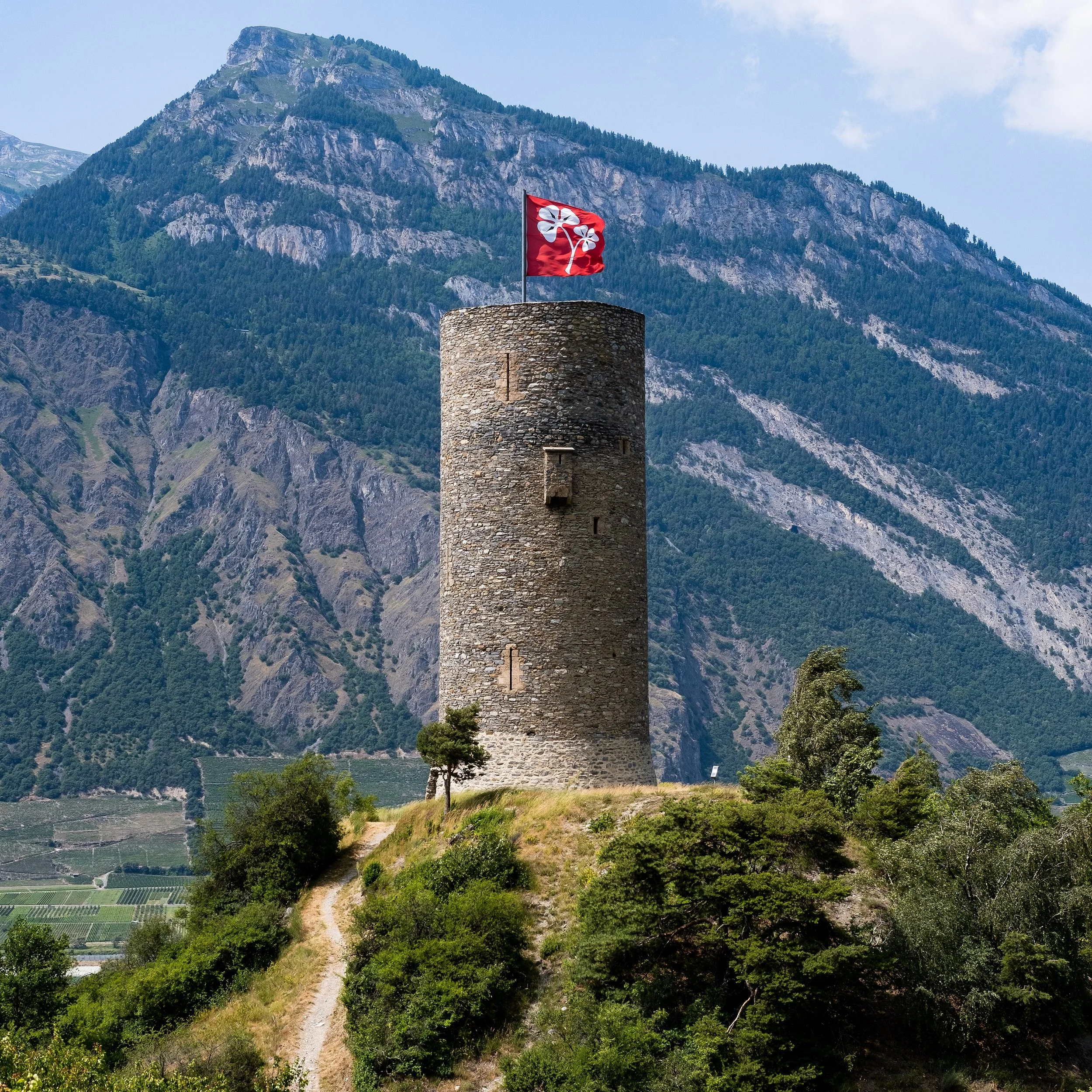 Une tour en pierre avec un drapeau rouge portant un symbole blanc, située dans un environnement montagneux avec une végétation verdoyante et des champs agricoles en contrebas.