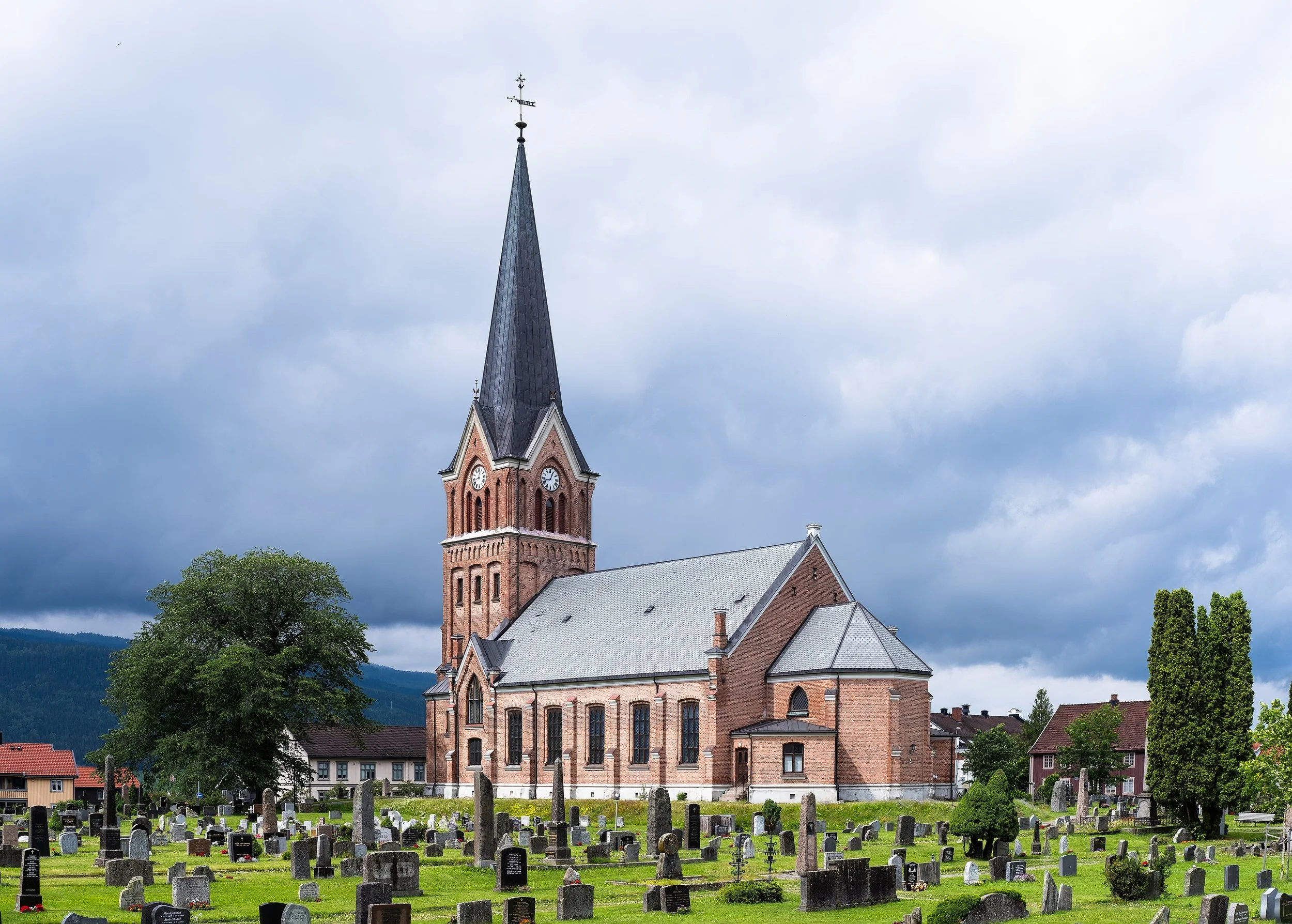 Une église en pierre rouge avec un clocher pointu et une horloge, entourée d'un cimetière avec des tombes et des arbres, sous un ciel nuageux.