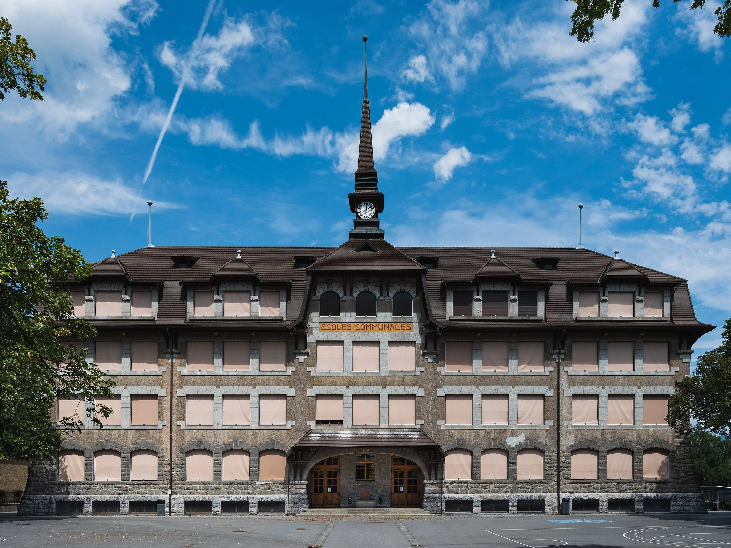 Bâtiment en pierre avec un toit en tuiles, une horloge au sommet, et une inscription 'Écoles Communales' sur la façade, sous un ciel bleu avec quelques nuages et un arbre à gauche.
