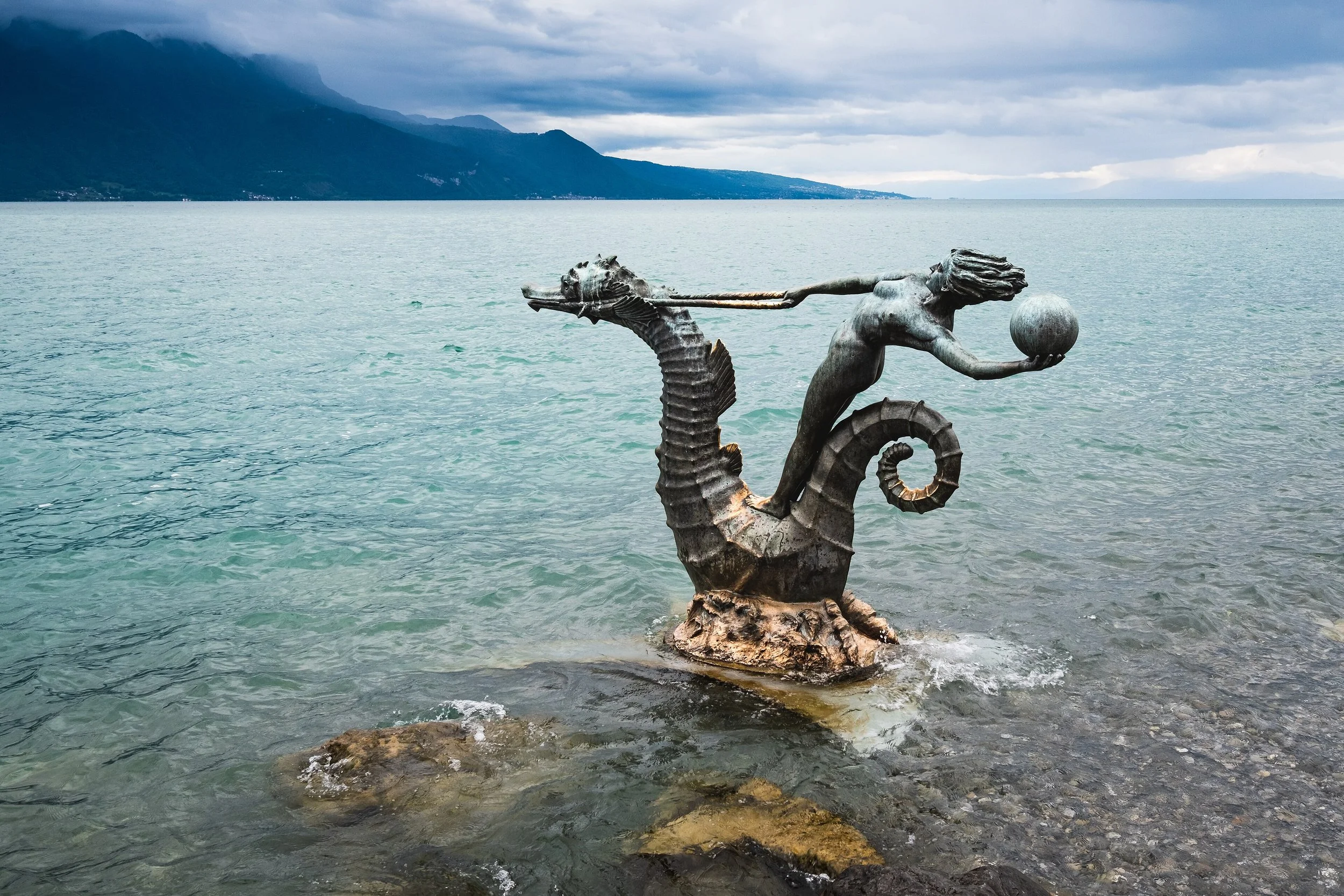 Statue en pierre représentant une figure humaine avec un globe, située dans un lac avec des montagnes en arrière-plan et un ciel nuageux.