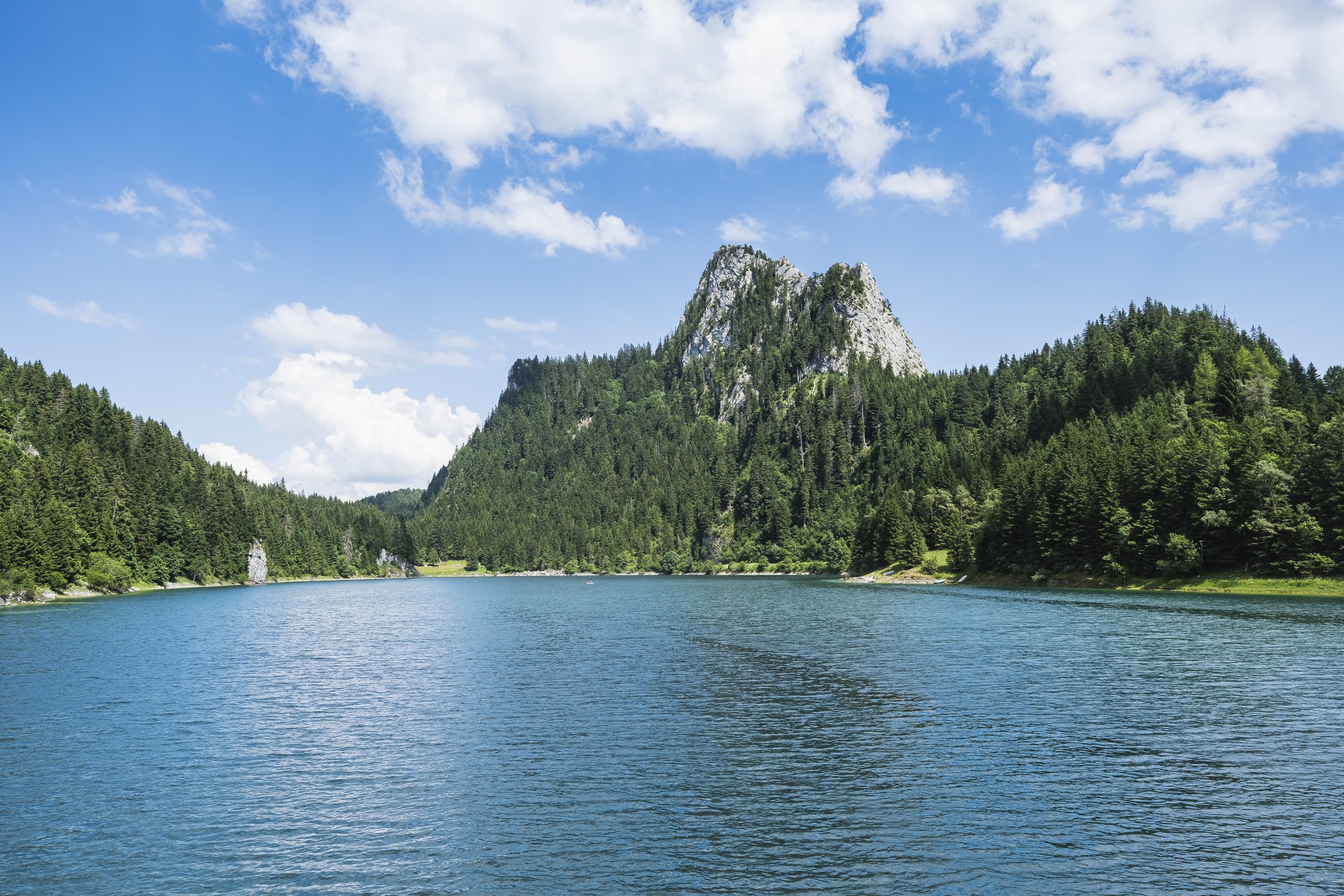 Un lac bleu entouré de forêts vertes avec une montagne rocheuse au centre, sous un ciel bleu avec quelques nuages.