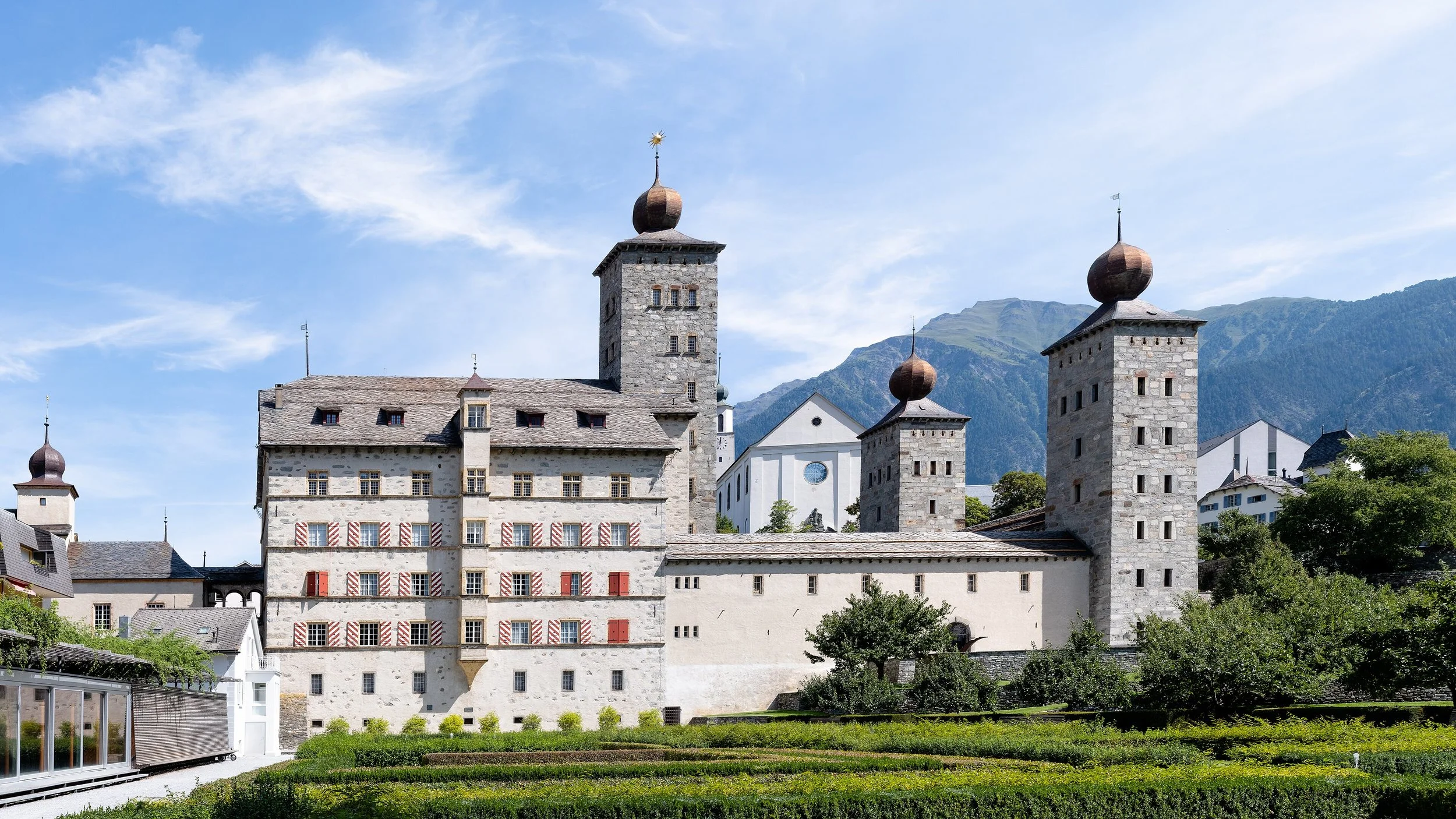 Château historique avec tours en pierre, située dans un paysage montagneux, sous un ciel bleu avec quelques nuages.