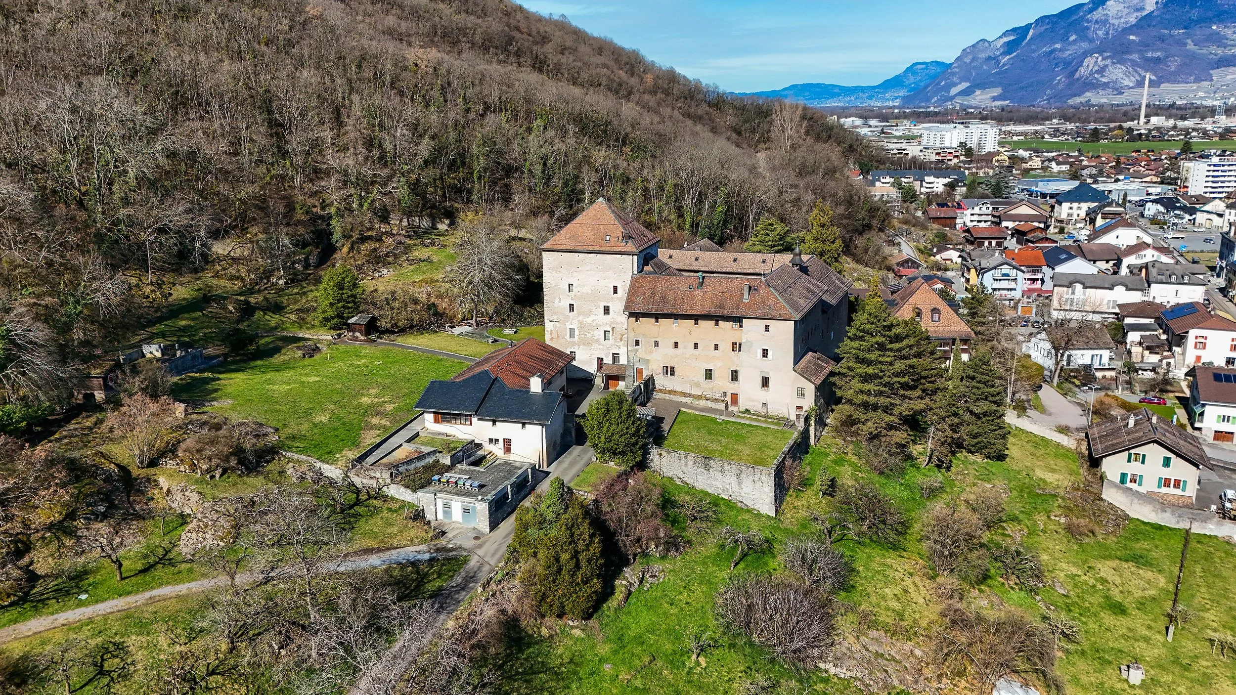 Photo aérienne d'un vieux château avec murs en pierre et toit en tuiles, entouré d'arbres et de jardins, avec un village et des montagnes en arrière-plan