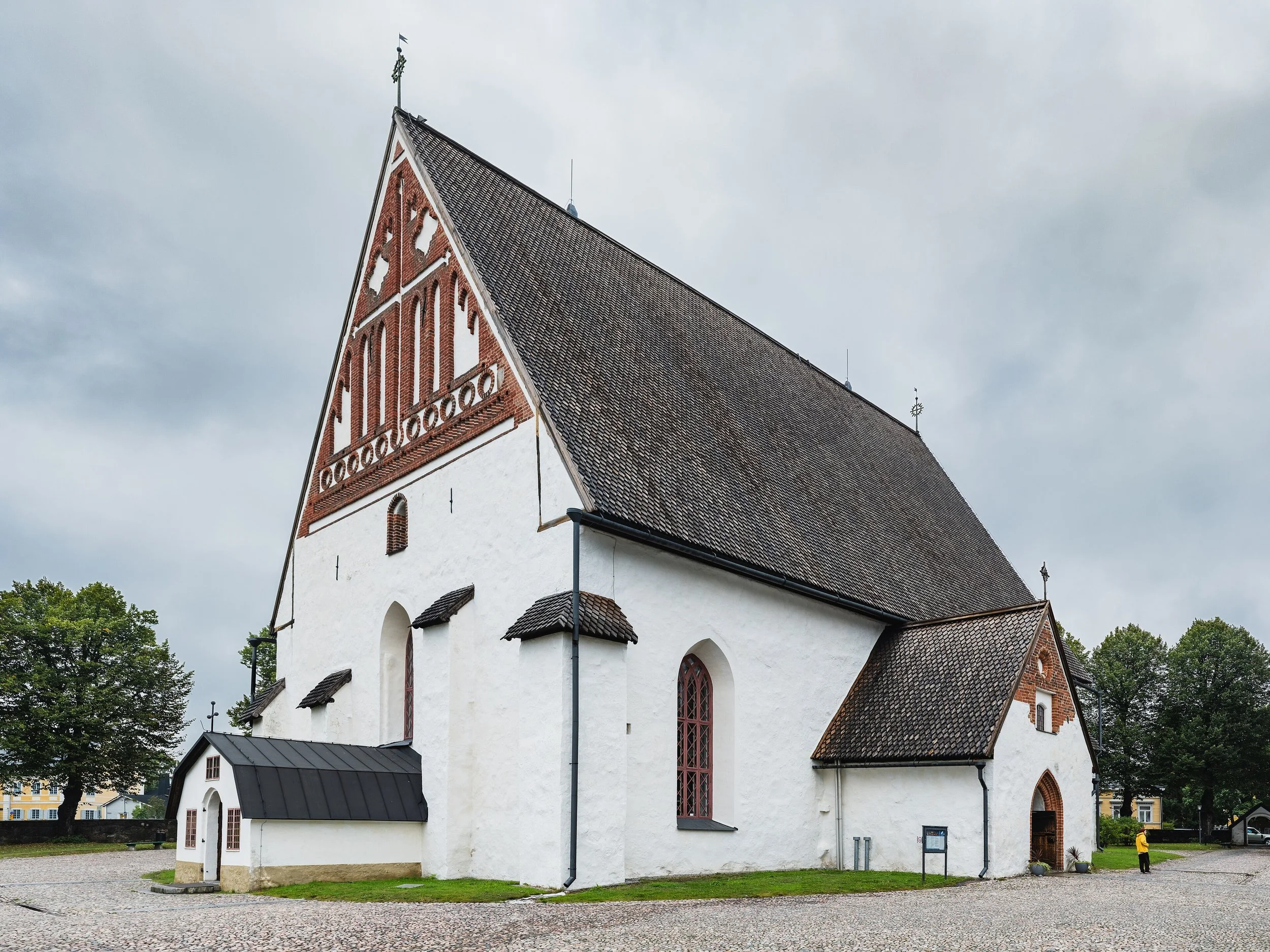 Une église en pierre blanche avec un toit en tuiles sombres et des fenêtres en arc, entourée d'arbres et d'une place pavée, par temps nuageux.
