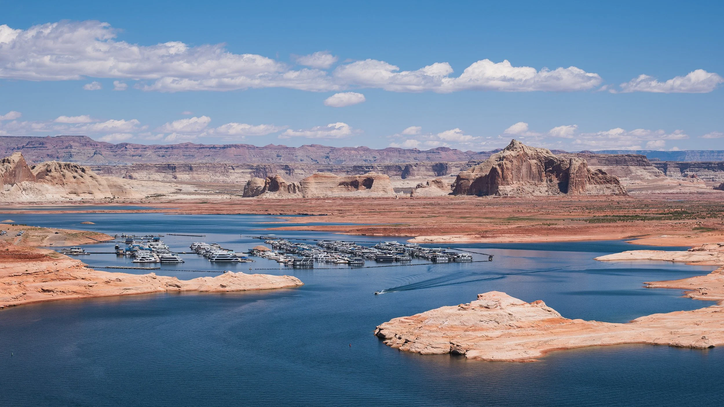 Vue panoramique du lac Powell avec des bateaux de plaisance amarrés, entouré de formations rocheuses rouges dans le canyon, sous un ciel bleu parsemé de nuages.