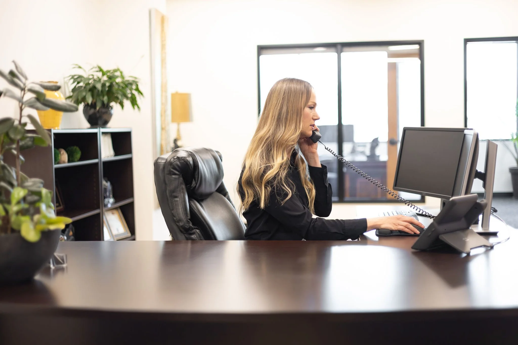 A woman with long blonde hair, wearing a black blazer, is sitting at a desk in an office, talking on a corded phone while working on a desktop computer. There are potted plants and shelves with decorative items in the background.
