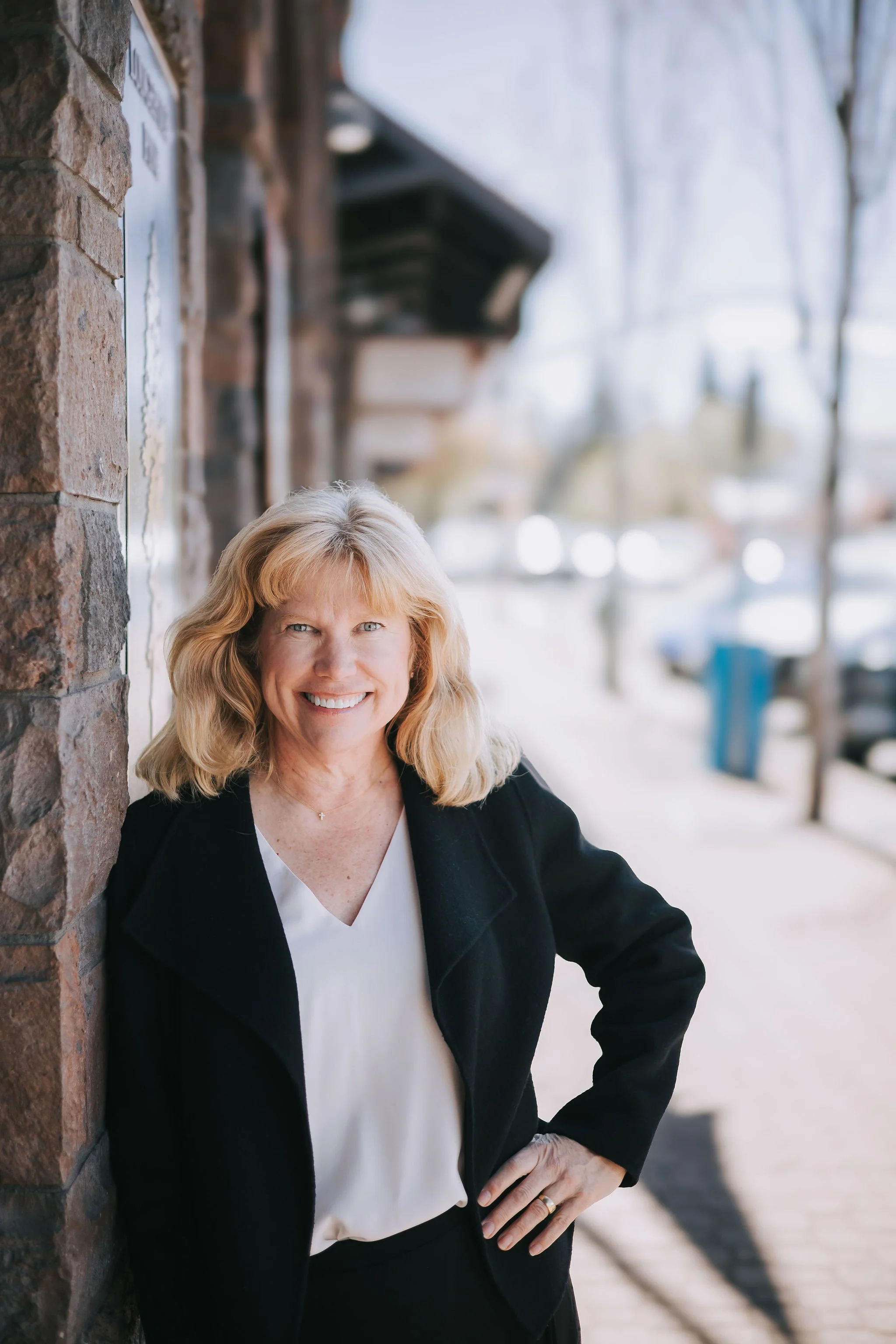 A smiling woman with blonde hair standing outdoors next to a brick wall, wearing a black blazer over a white blouse, with one hand on her hip.