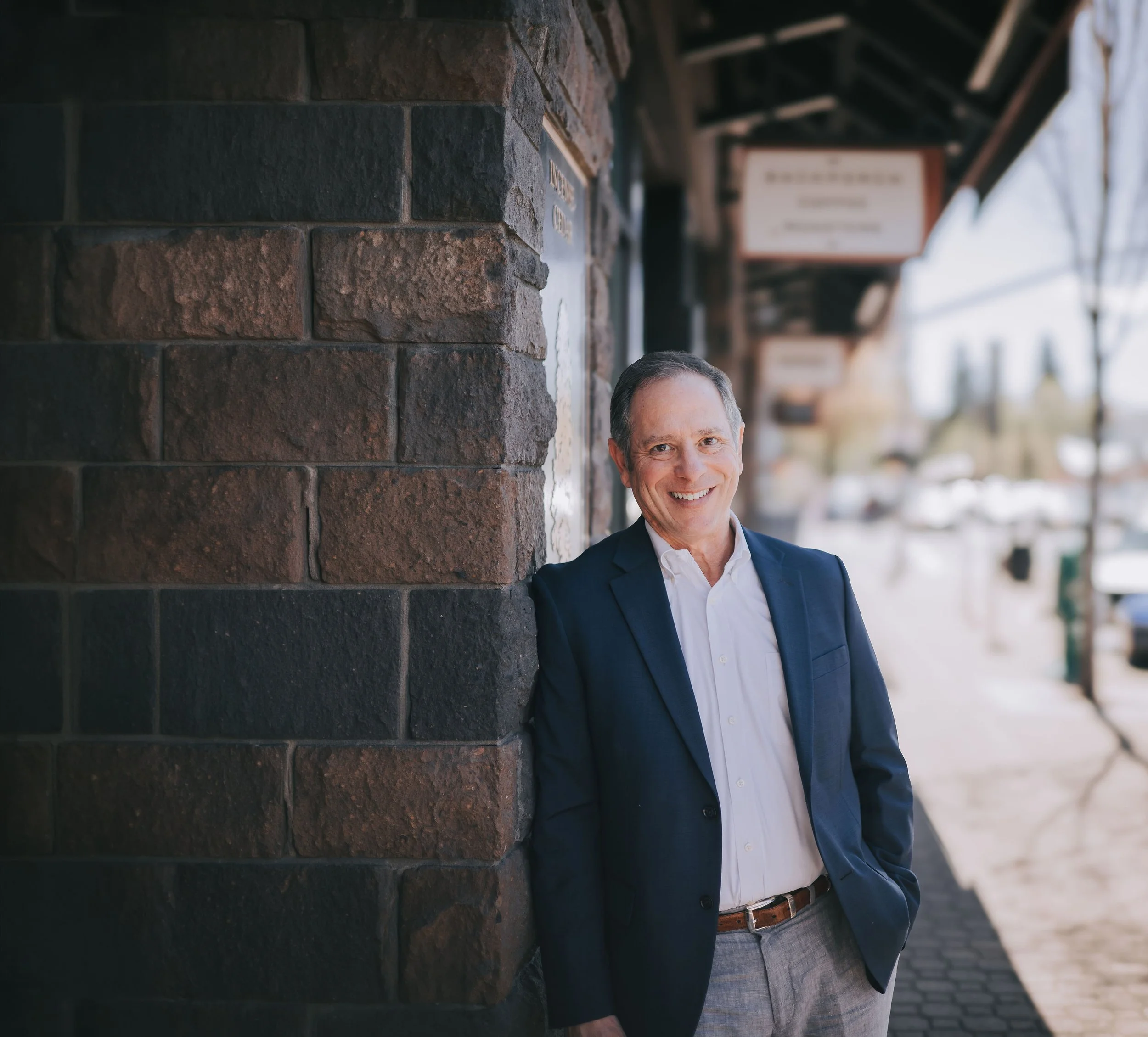 A man in a business suit standing outdoors, smiling, with a brick building and signs in the background.