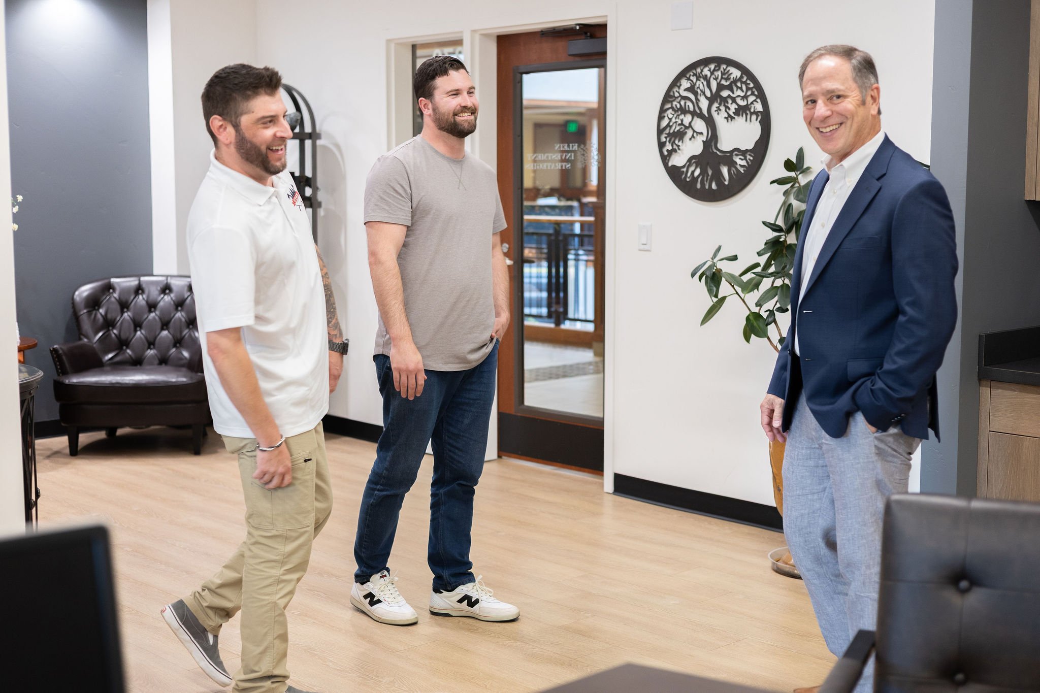 Three men are smiling and talking indoors, standing casually in front of a white wall with a black tree wall art and a potted plant. One man is wearing a white polo shirt and beige pants, another is in a gray t-shirt and jeans, and the third is dress