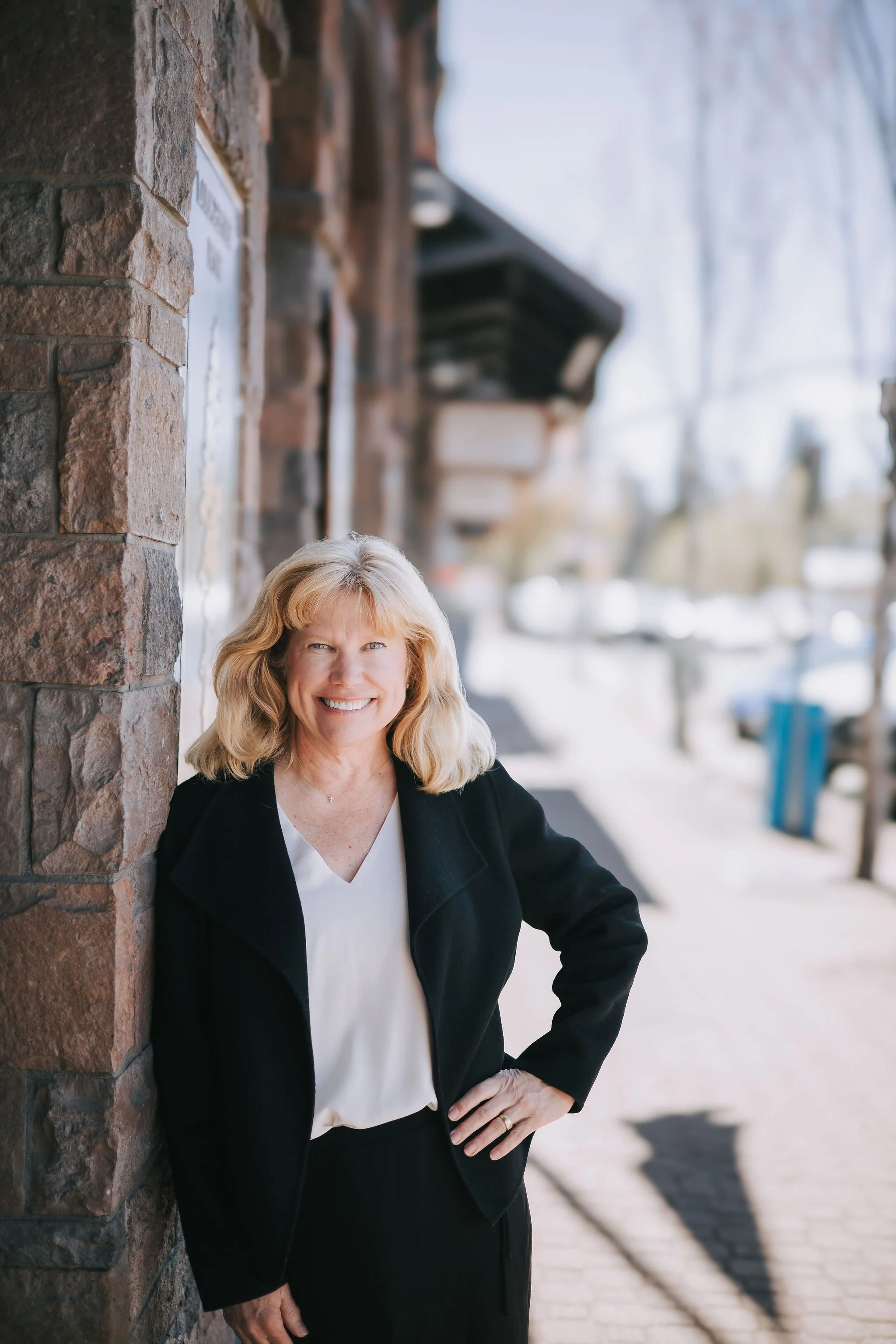 A smiling woman with blonde hair, wearing a black blazer and white blouse, standing outdoors near a brick wall on a sunny day.