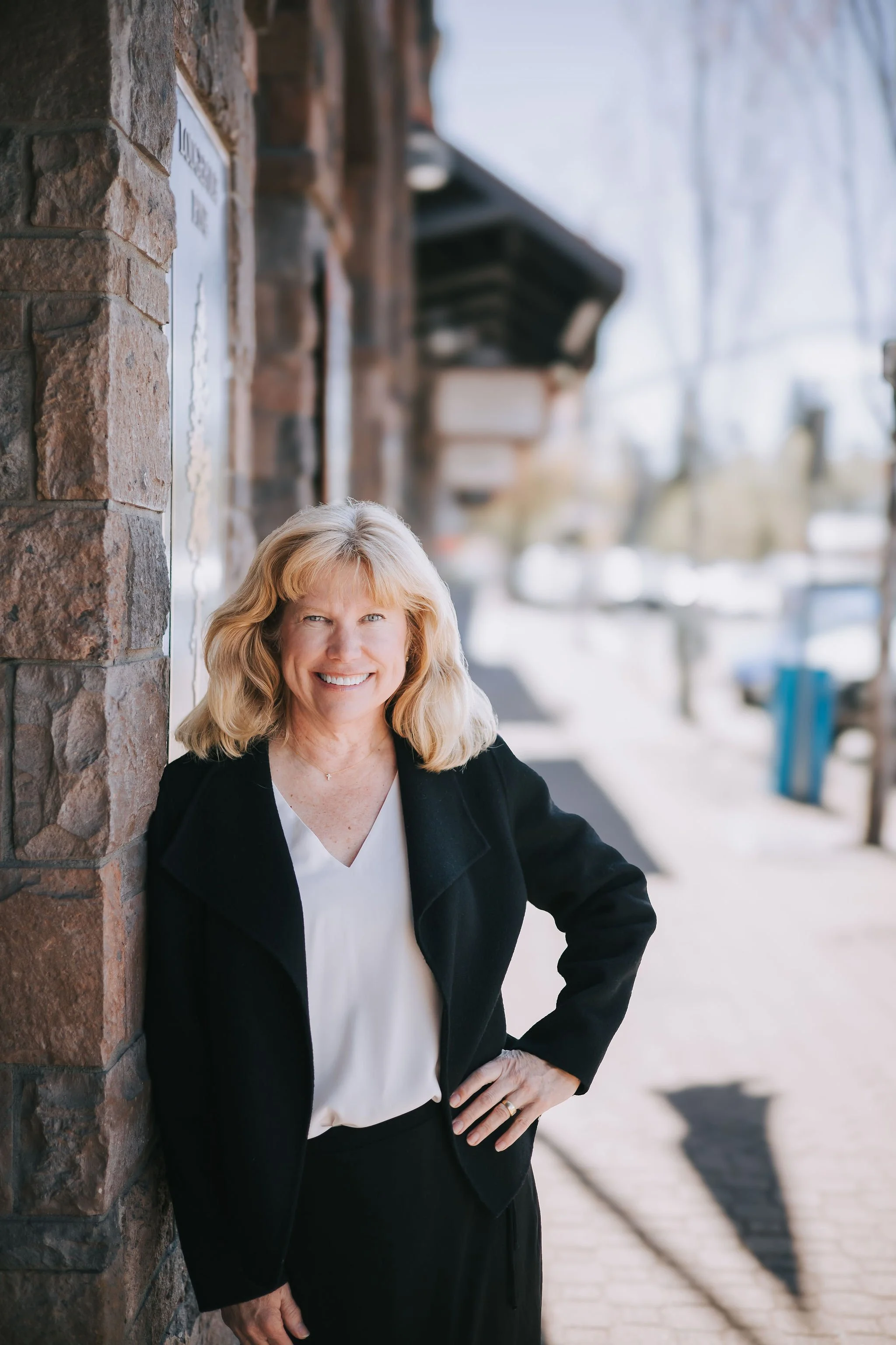 A woman with blonde hair smiling, dressed in a black blazer and white top, standing outdoors beside a brick wall, with a blurred background of a sidewalk and buildings.