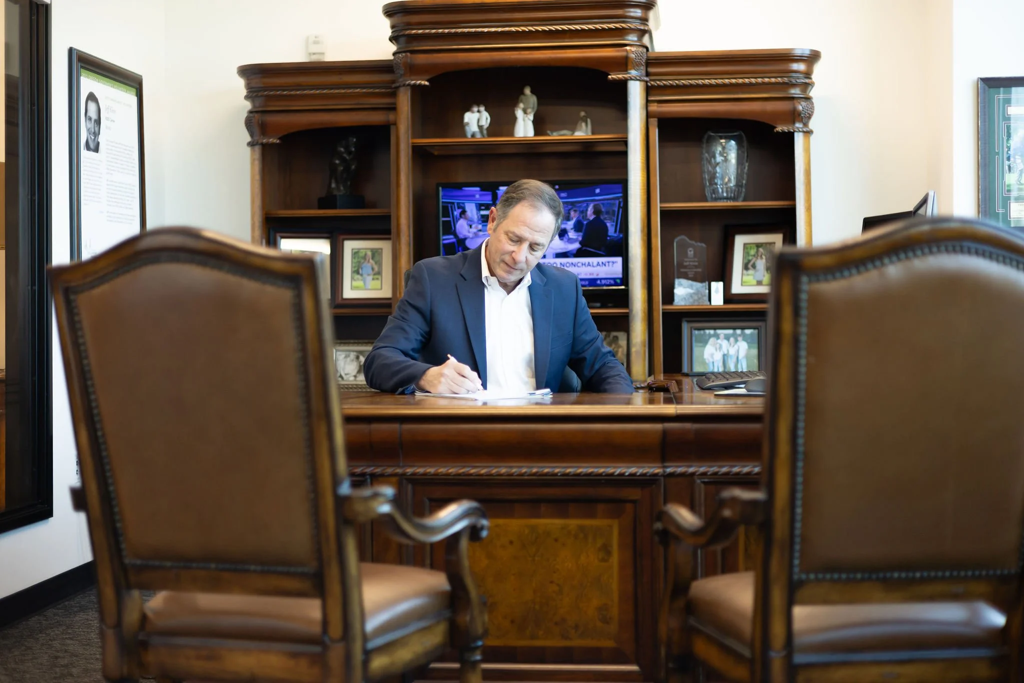 Jeff Klein of Klein Investment Strategies, financial investment firm located in Bend, OR. Office with wooden shelves, framed photos, and a television in the background. The photo is taken through a gap between two chairs.
