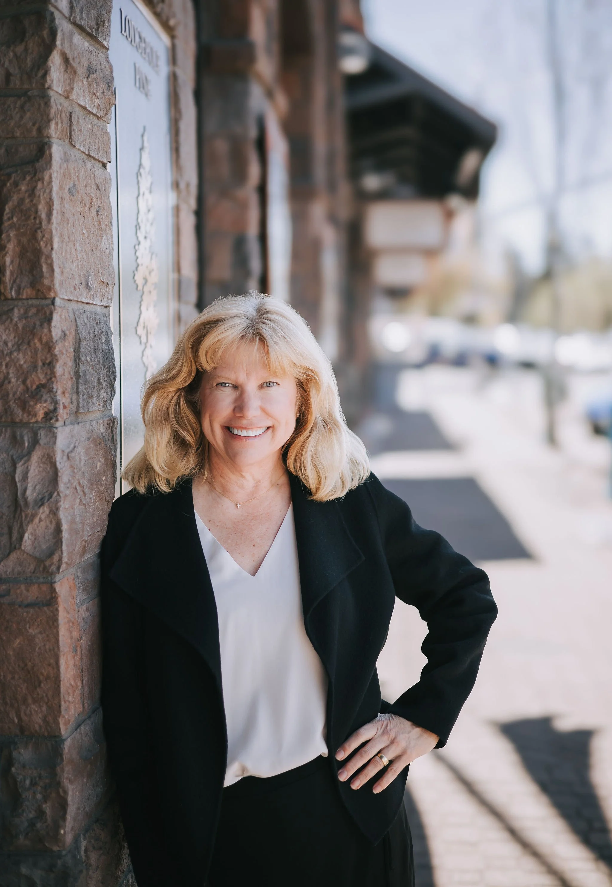 A smiling middle-aged woman with blonde hair, wearing a black blazer and white blouse, standing outdoors against a brick wall with blurred background.