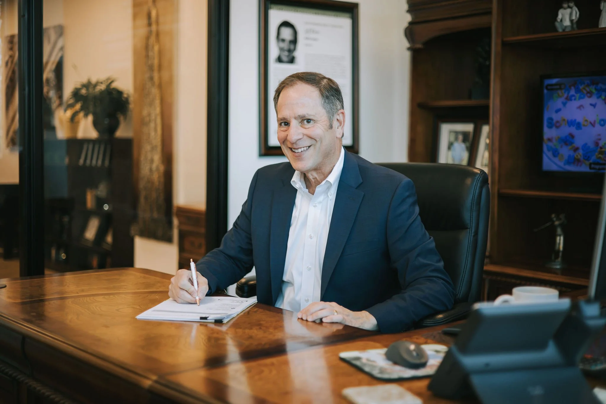 Jeff Klein of Klein Investment Strategies, financial investment firm located in Bend, OR holding a pen over a stack of papers, in an office with wooden shelves, framed photos, and a framed portrait in the background.