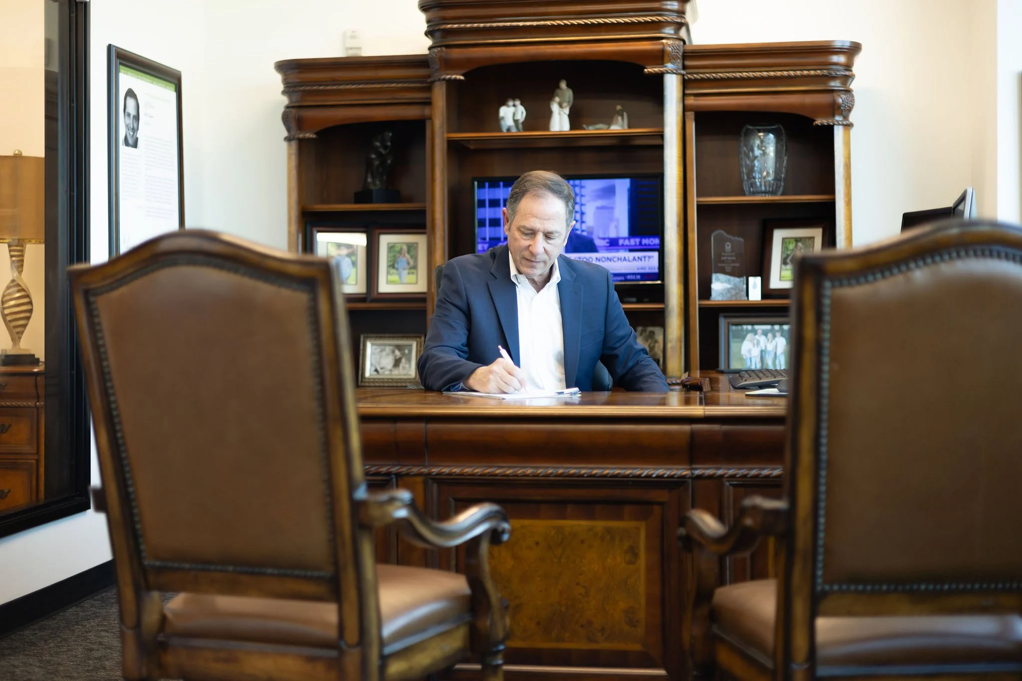 Jeff Klein of Klein Investment Strategies, financial inve in a suit sitting at a large wooden desk writing on paper with a pen inside an office. Behind him is a wooden bookshelf with framed photos, decorative items, and a TV showing a news broadcast.