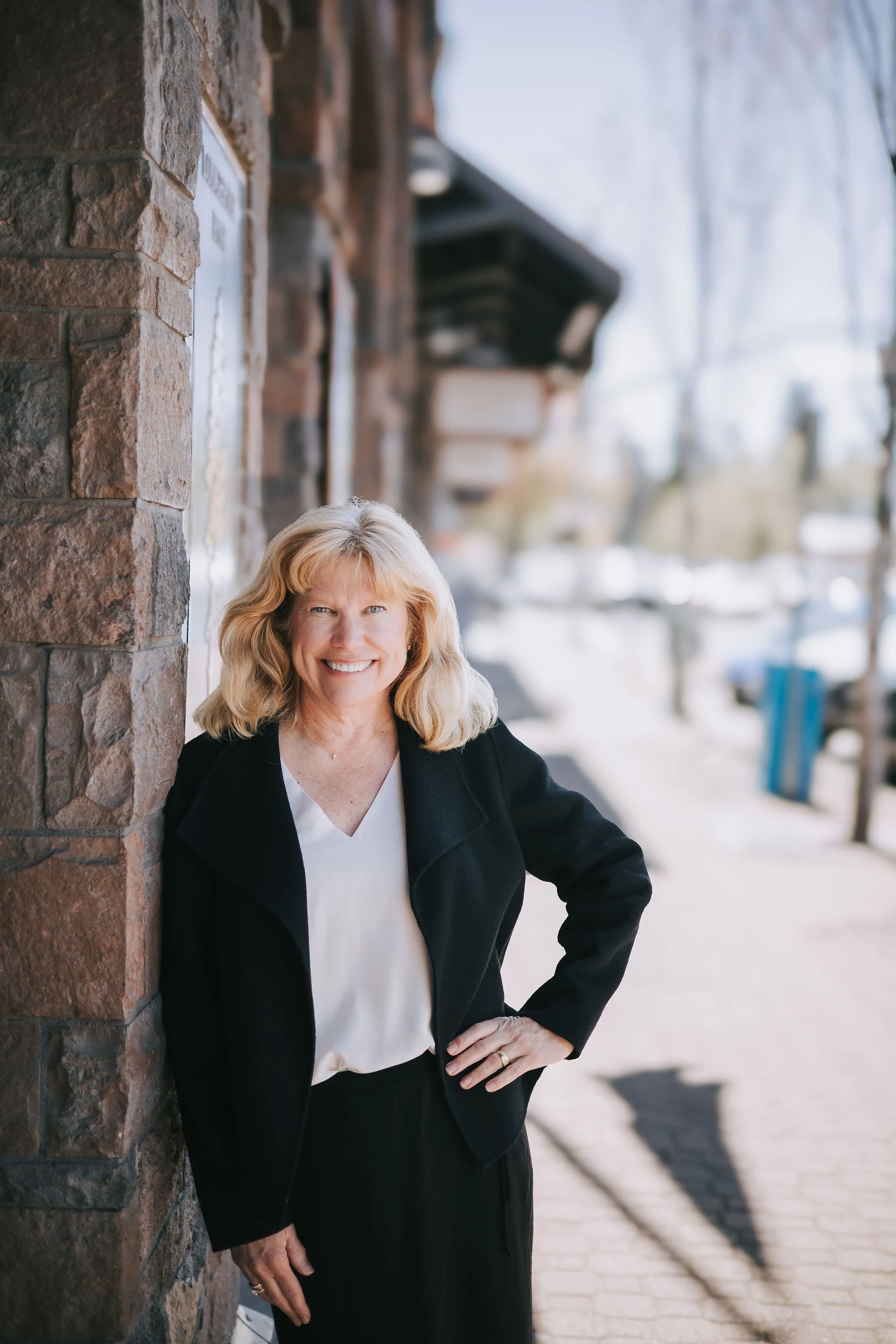 A smiling middle-aged woman with blonde hair standing outdoors against a brick wall. She is wearing a black blazer over a white top, with her left hand on her hip. The background features a blurred street scene with leafless trees and a blue bench.