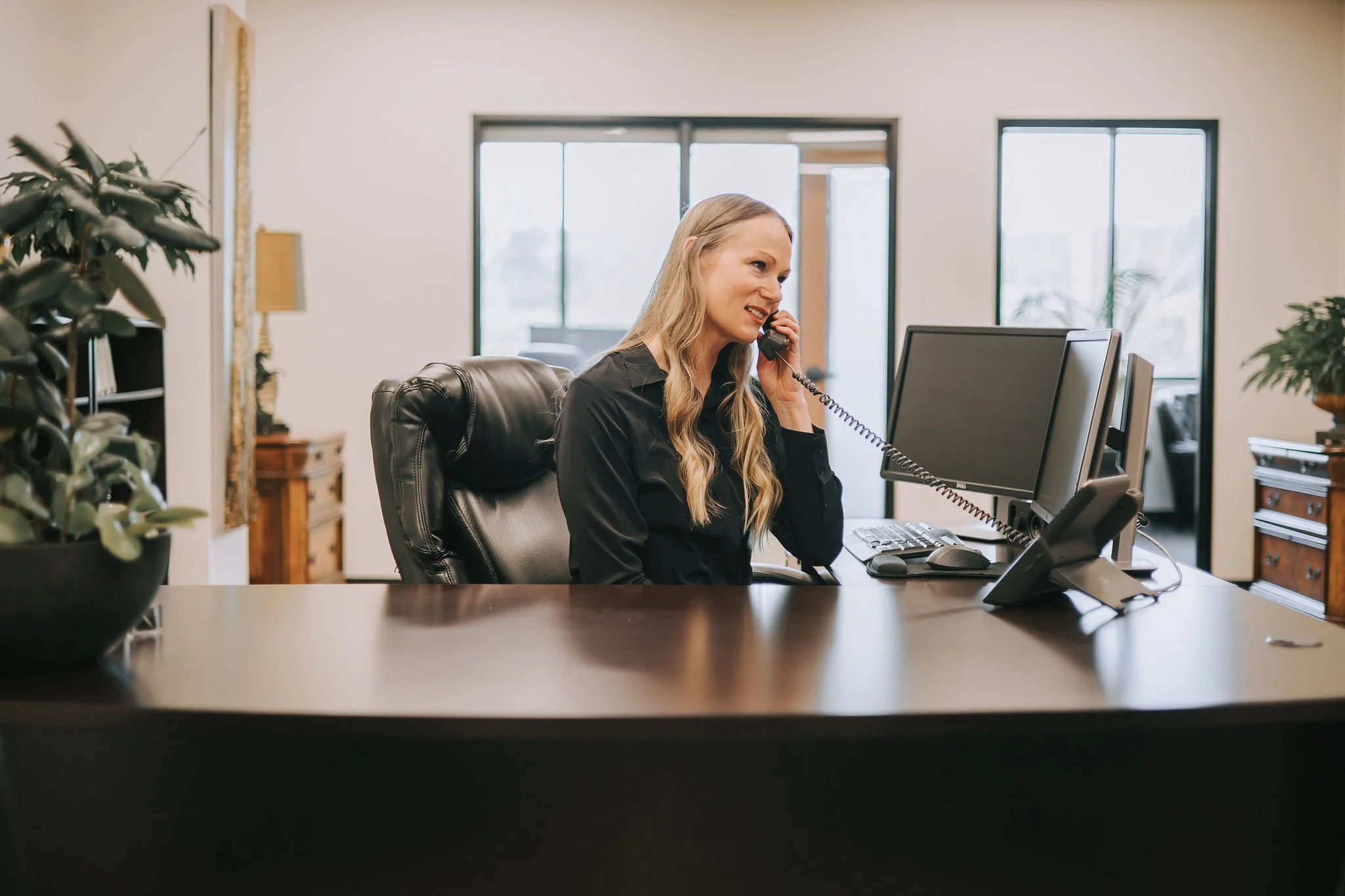 A woman with long blonde hair, wearing a black blouse, talking on a landline phone at her office desk with two computer monitors, a keyboard, and a mouse. There are large windows and indoor plants in the background.
