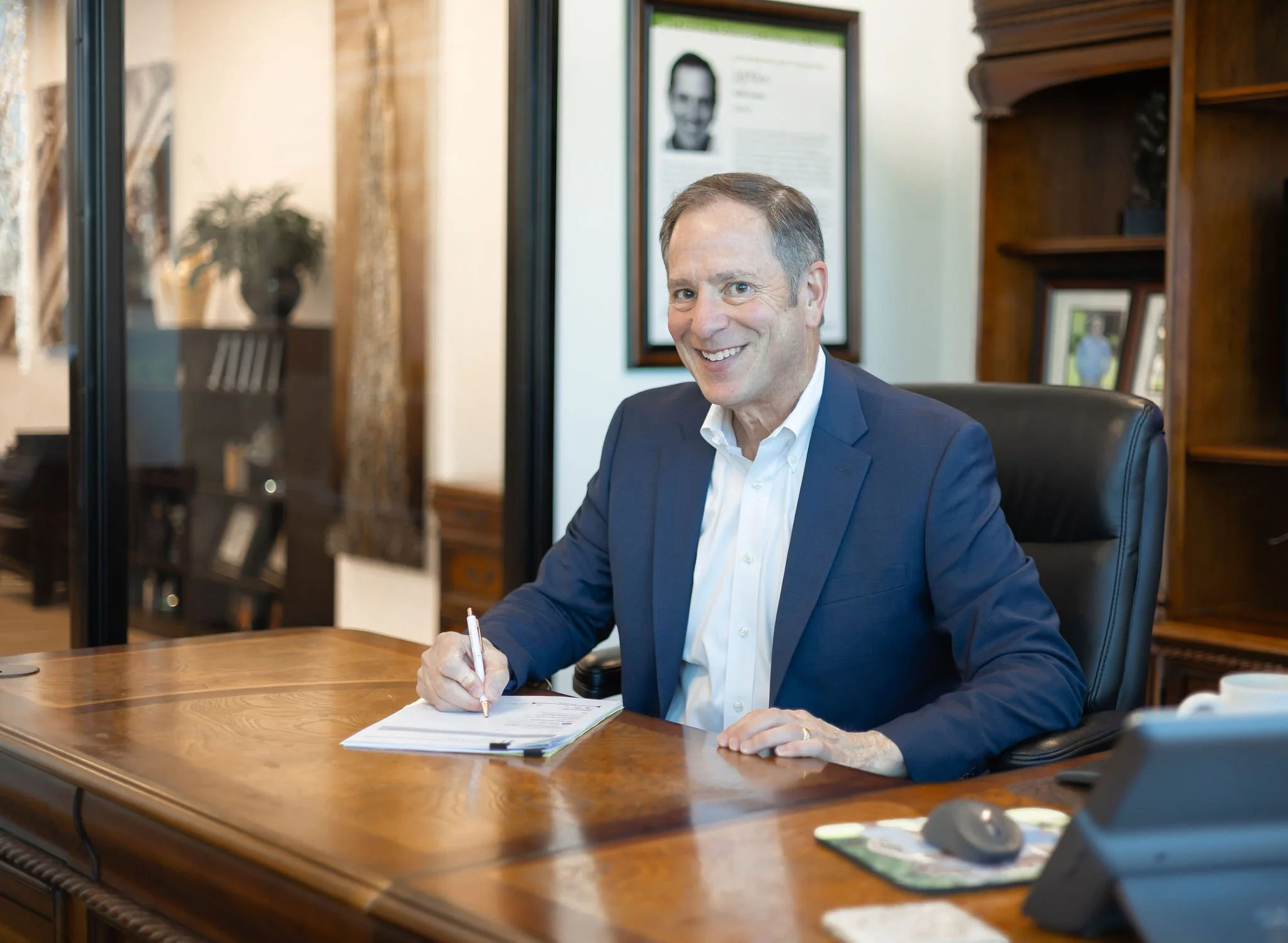Jeff Klein of Klein Investment Strategies, financial investment firm located in Bend, OR holding a pen over a stack of papers, in an office with wooden shelves, framed photos, and a framed portrait in the background.