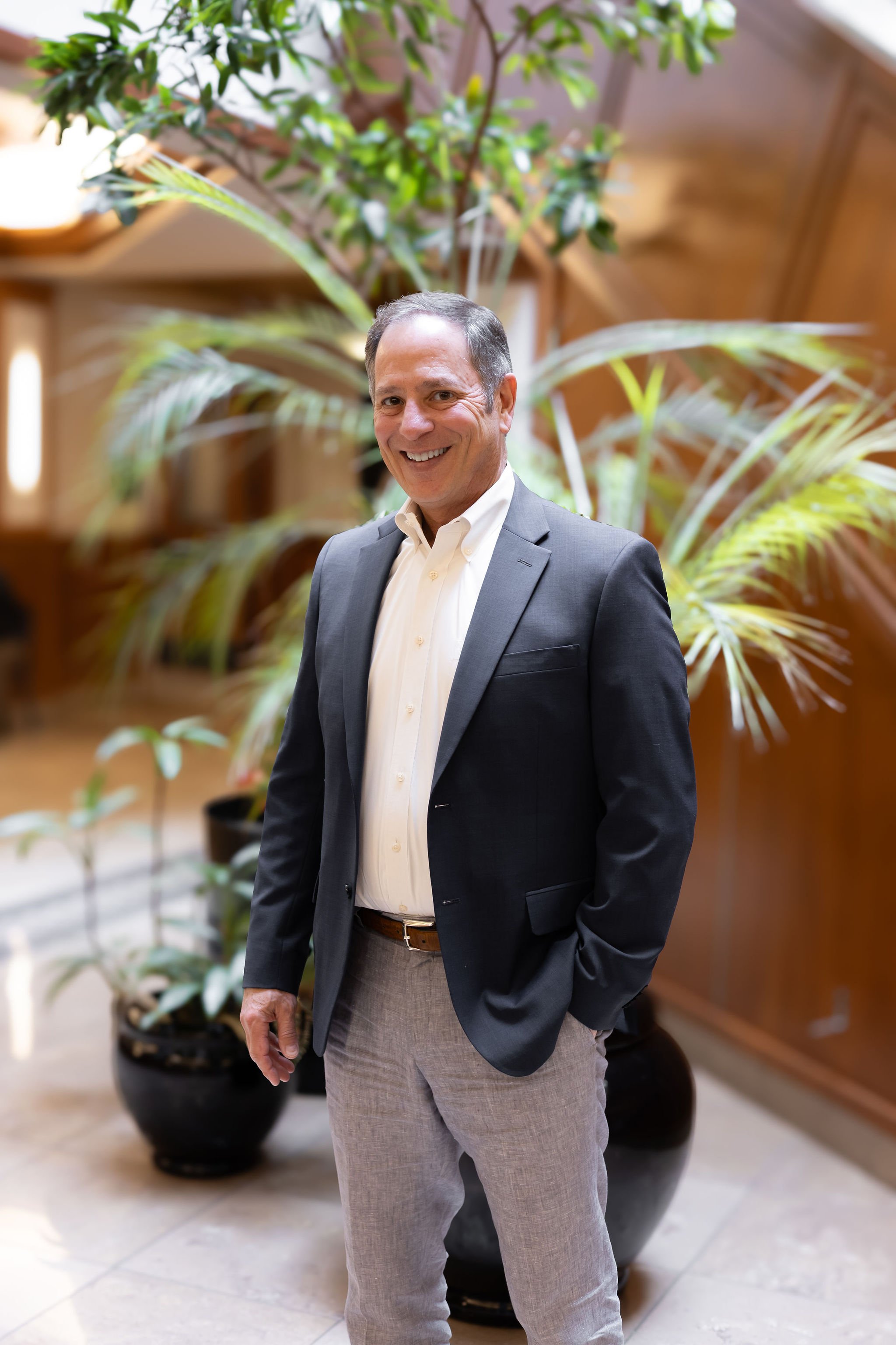 A smiling Jeff Klein of Klein Investment Strategies, an Oregon financial investment firm, in a gray suit jacket, white shirt, and light gray pants standing indoors in front of a large potted plant with green leaves, wooden walls, and a tiled floor.