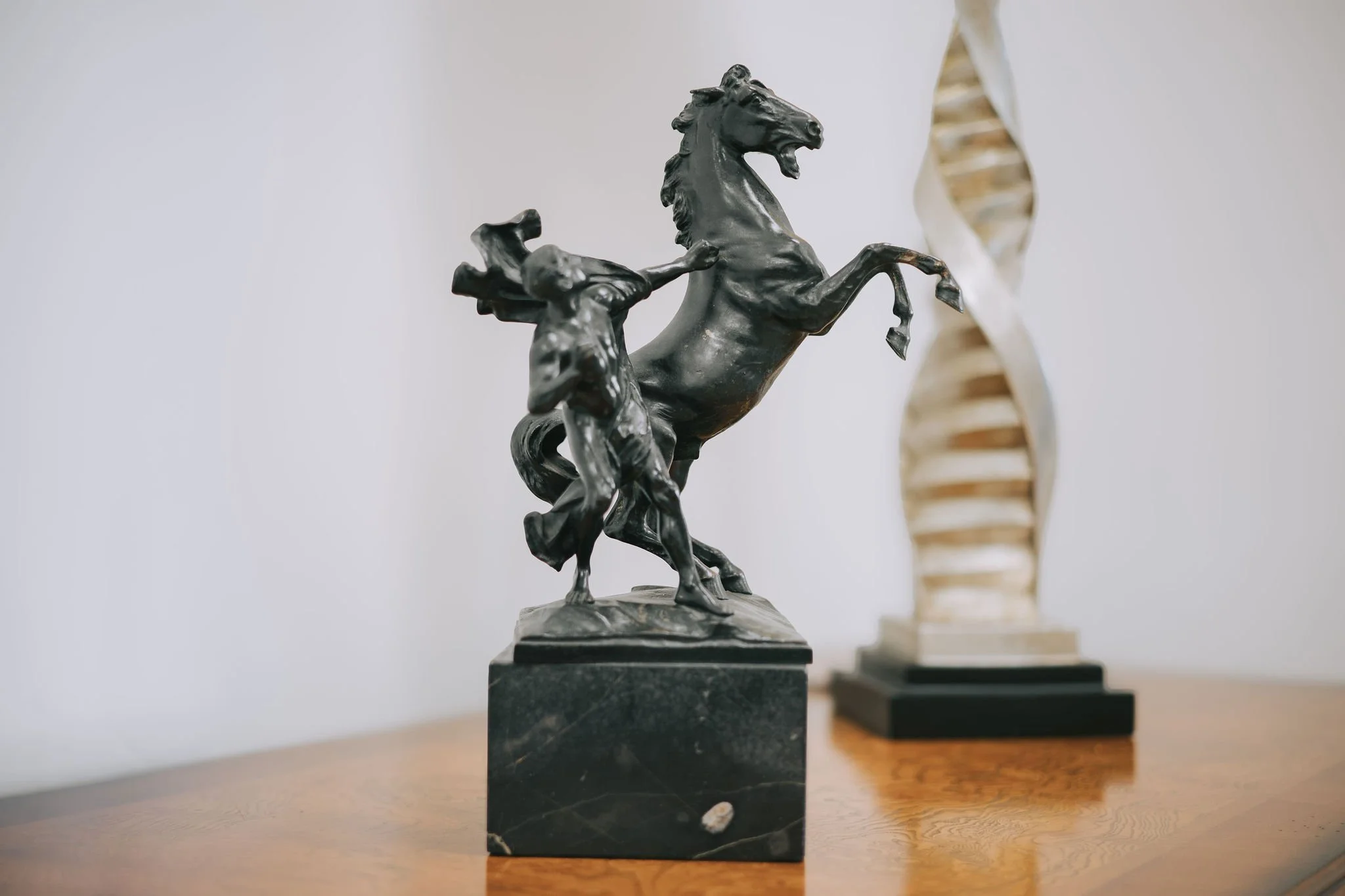 A black metal sculpture of a horse rearing up, placed on a black marble base, on a wooden surface with two other sculptures visible in the background.