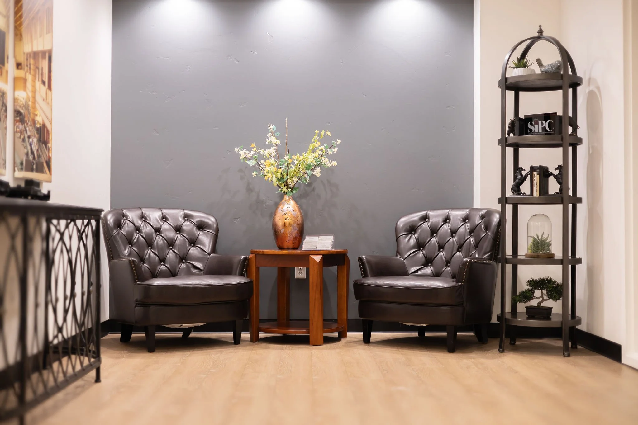 Interior of a waiting or lounge area with two black leather tufted chairs, a small wooden table with a decorative vase with flowers, a gray accent wall, and a black shelving unit with decorative items and books.