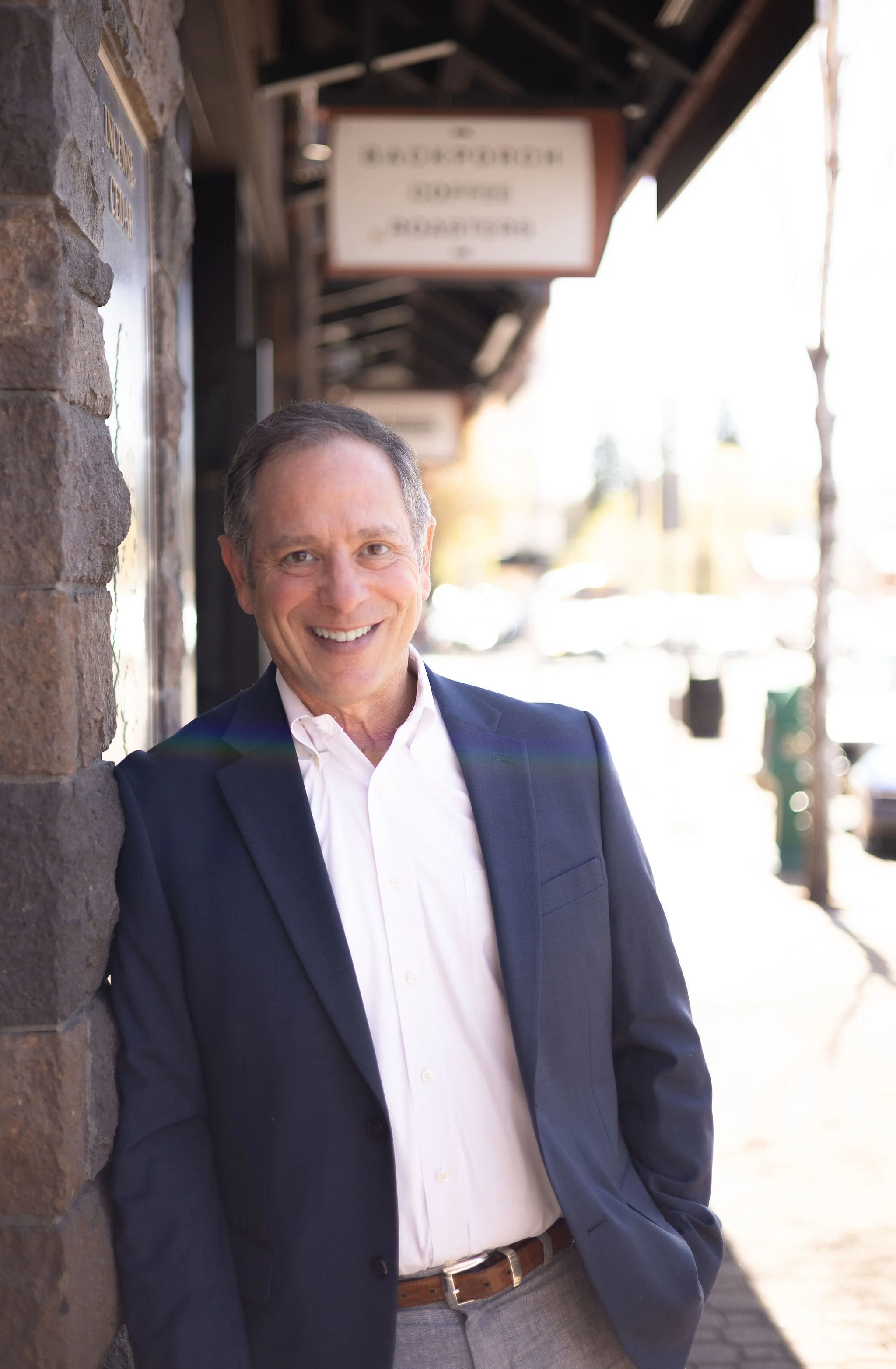 A man in a navy blazer and white shirt smiling and leaning against a brick wall on a sidewalk.