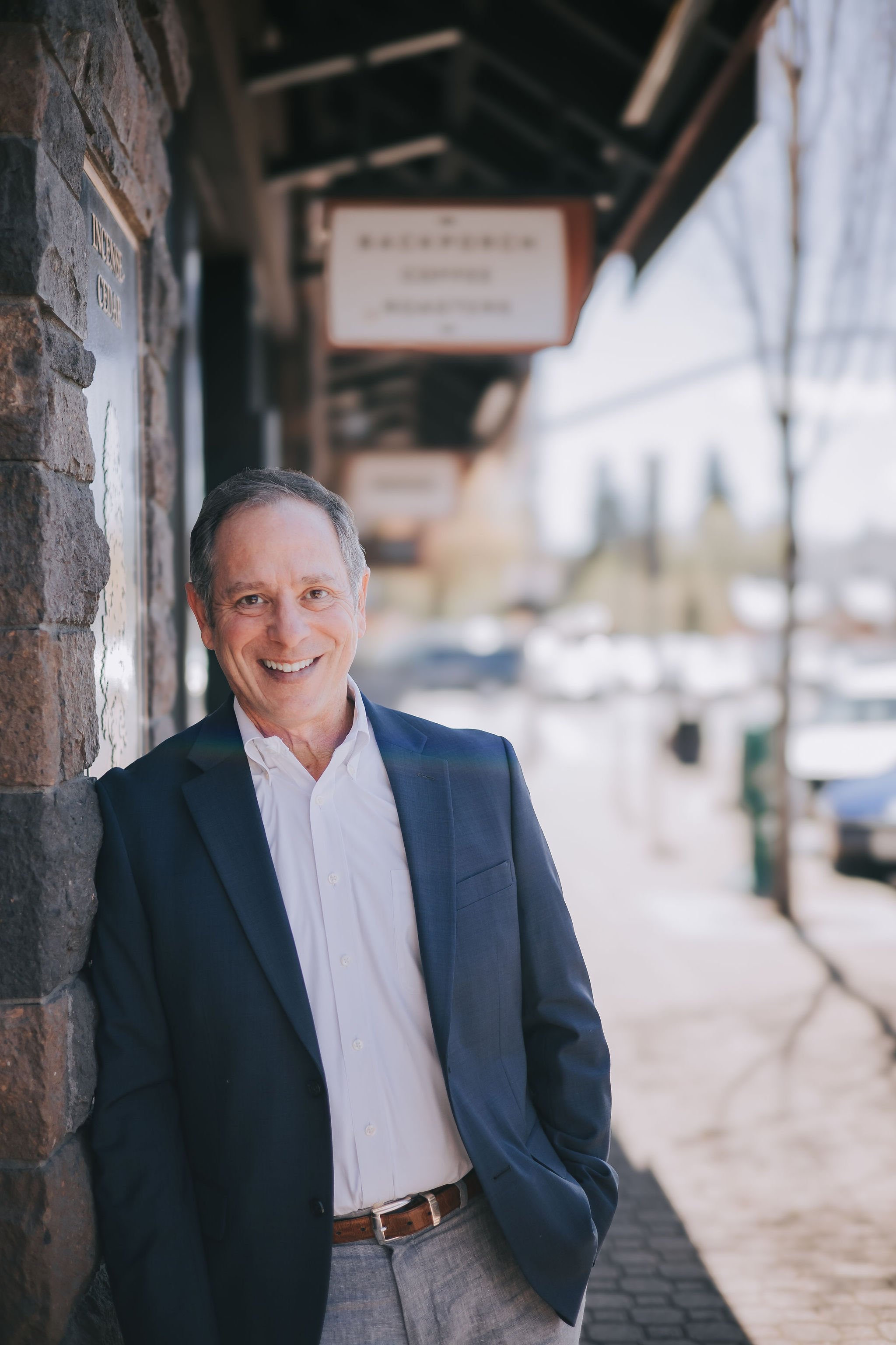 A man in a suit jacket and dress shirt smiling and standing outdoors, partially leaning against a brick wall, with a blurred street scene in the background.
