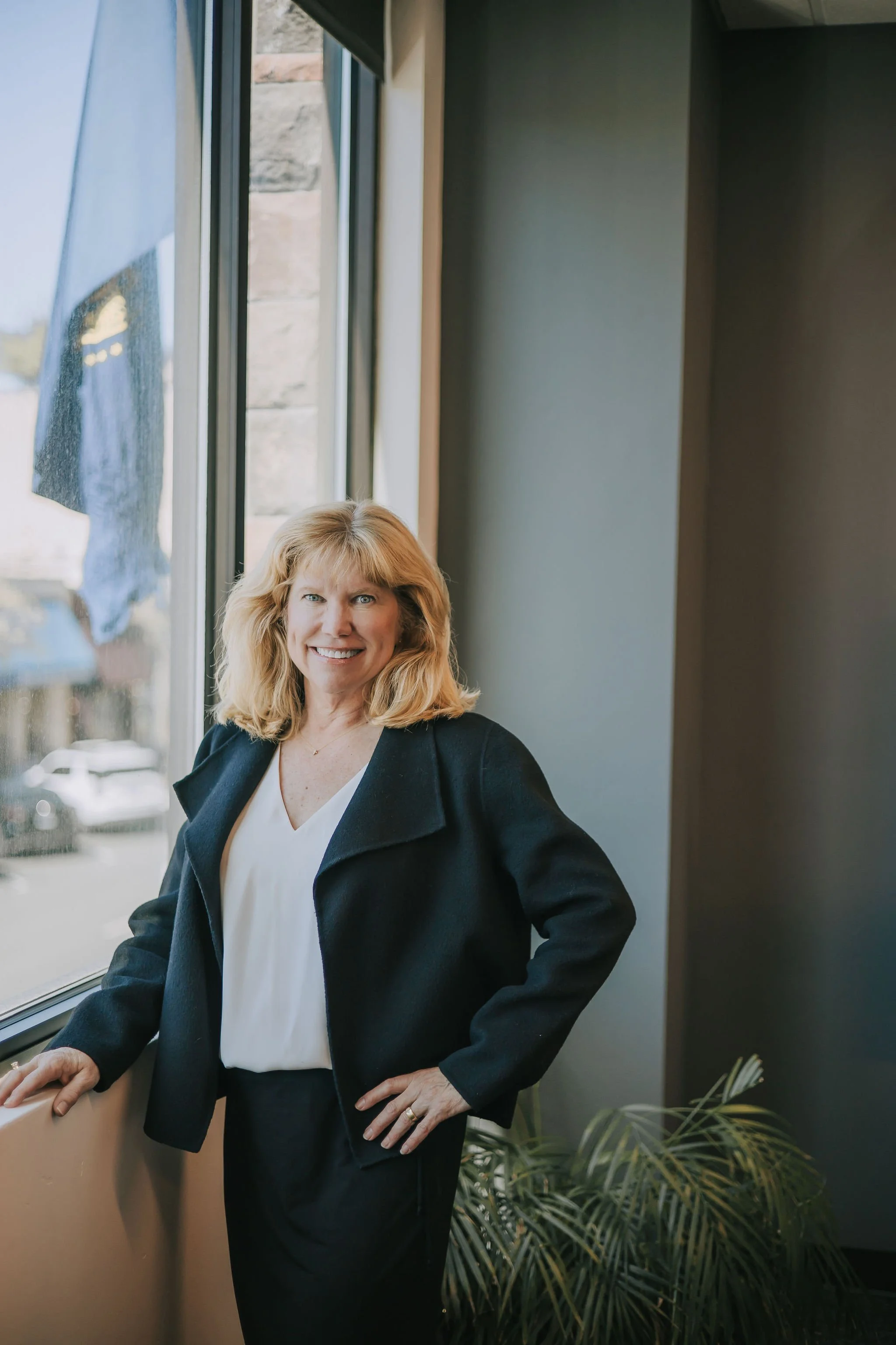 A smiling Michelle Klein of Klein Investment Strategies, an Oregon financial investment firm, with blonde hair wearing a white blouse and black blazer inside a building with large windows and a city view outside.