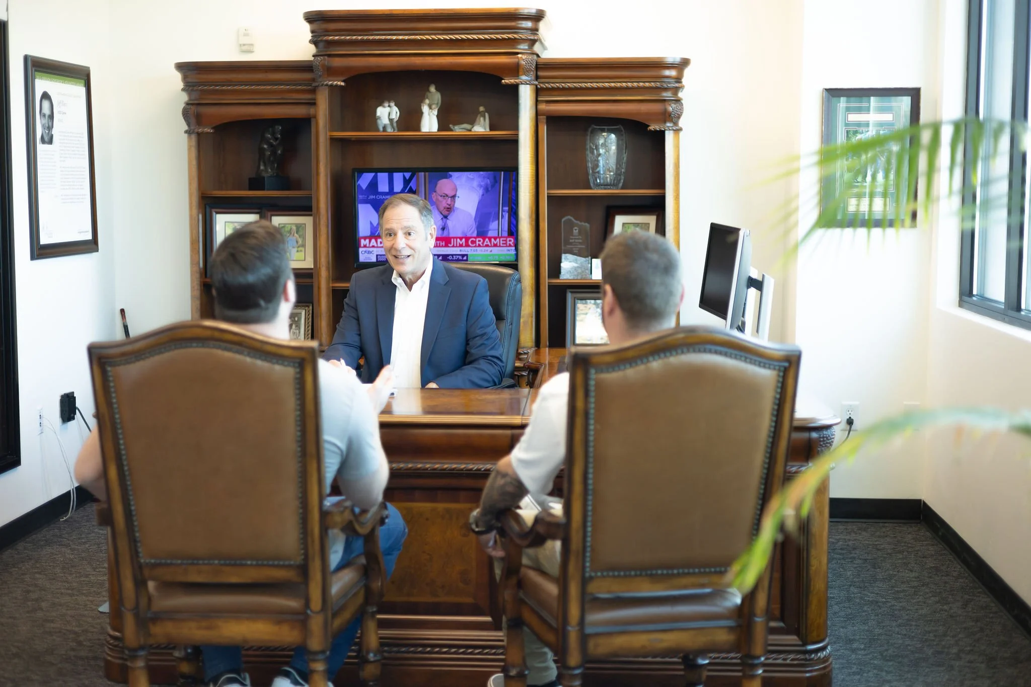 A man in a suit sitting at a wooden desk during a business meeting with two other people, with a TV showing a news program, framed artwork and a decorative shelf with sculptures in the background.