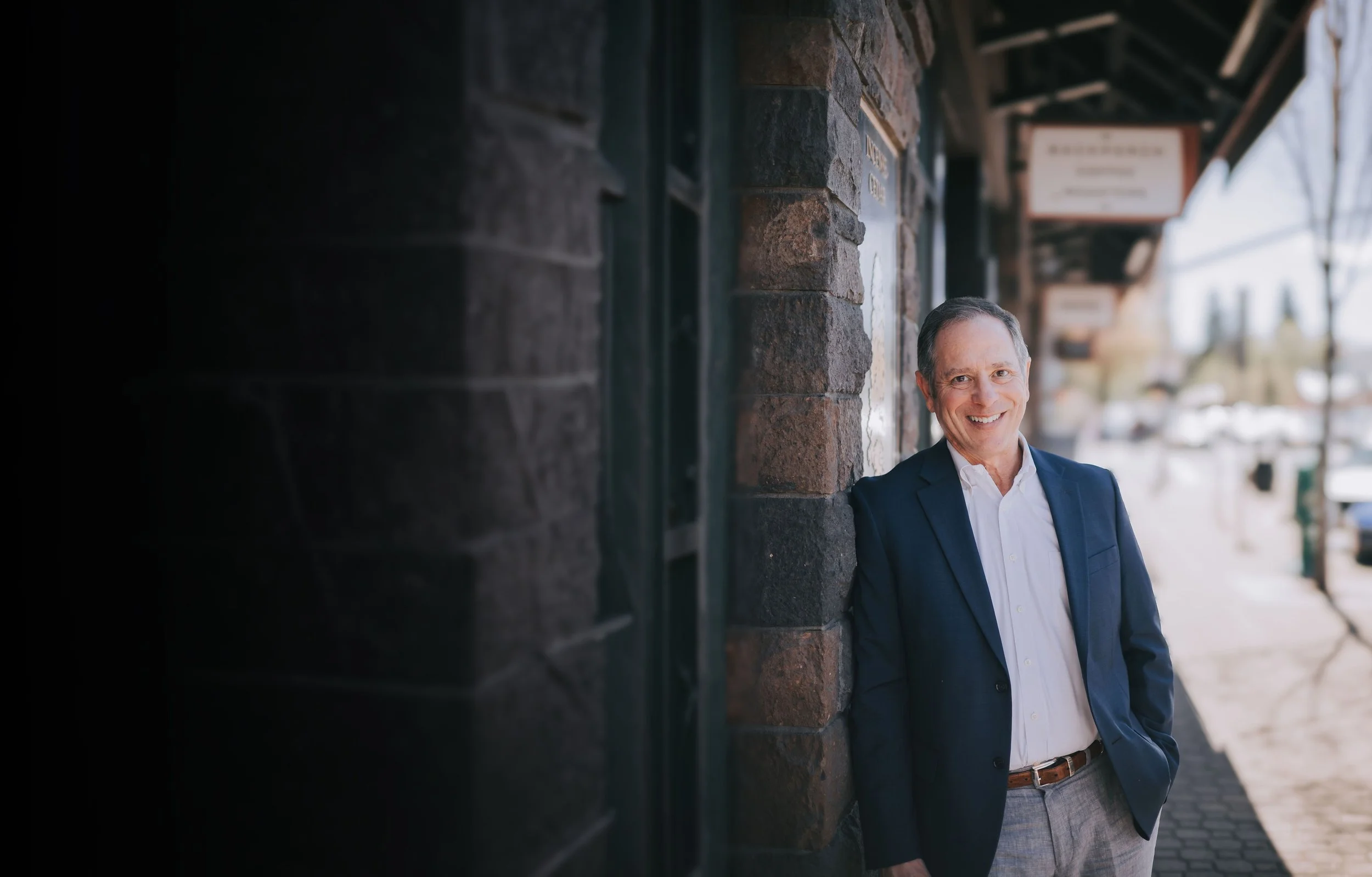 A man in a navy blazer and white shirt leaning against a brick wall on a sidewalk, smiling at the camera with blurred storefronts and trees in the background.
